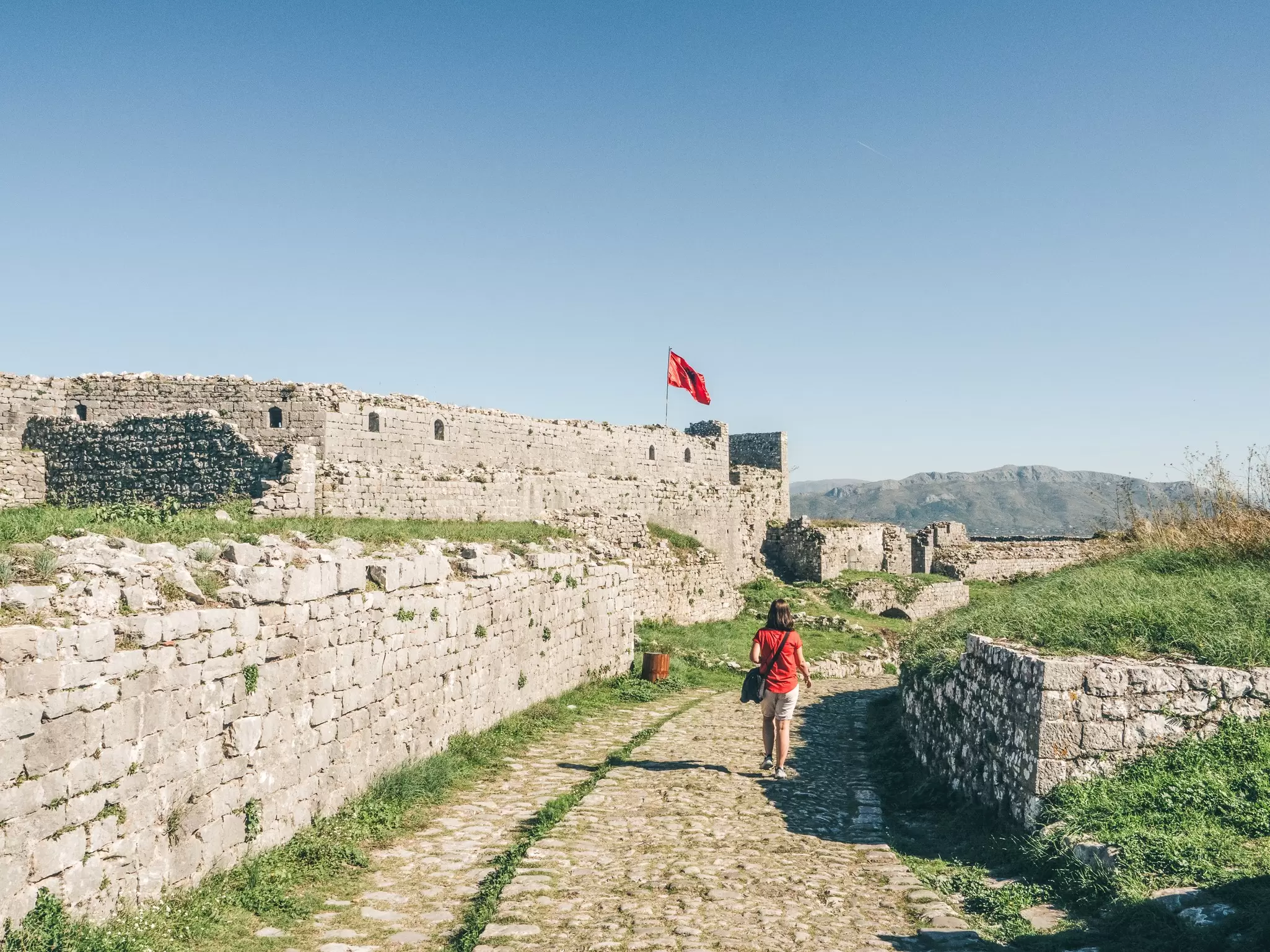 A woman walks towards a vast fortress with an Albanina flag flying above it: the Rozafa Castle near the city of Shkodër, Albania.