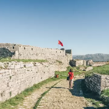 A woman walks towards a vast fortress with an Albanina flag flying above it: the Rozafa Castle near the city of Shkodër, Albania.