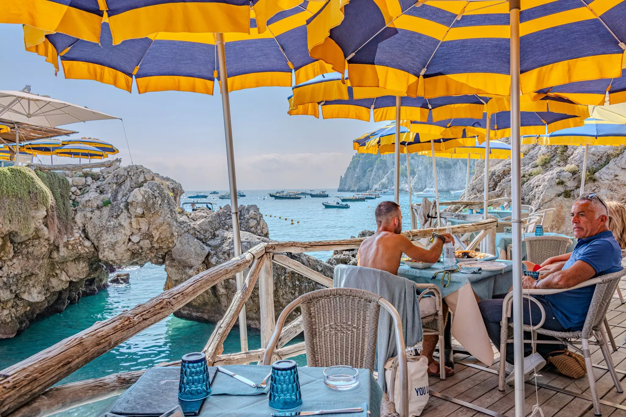 Two men sit a table on an outdoor terrace overlooking a cliff-Ringed cove.