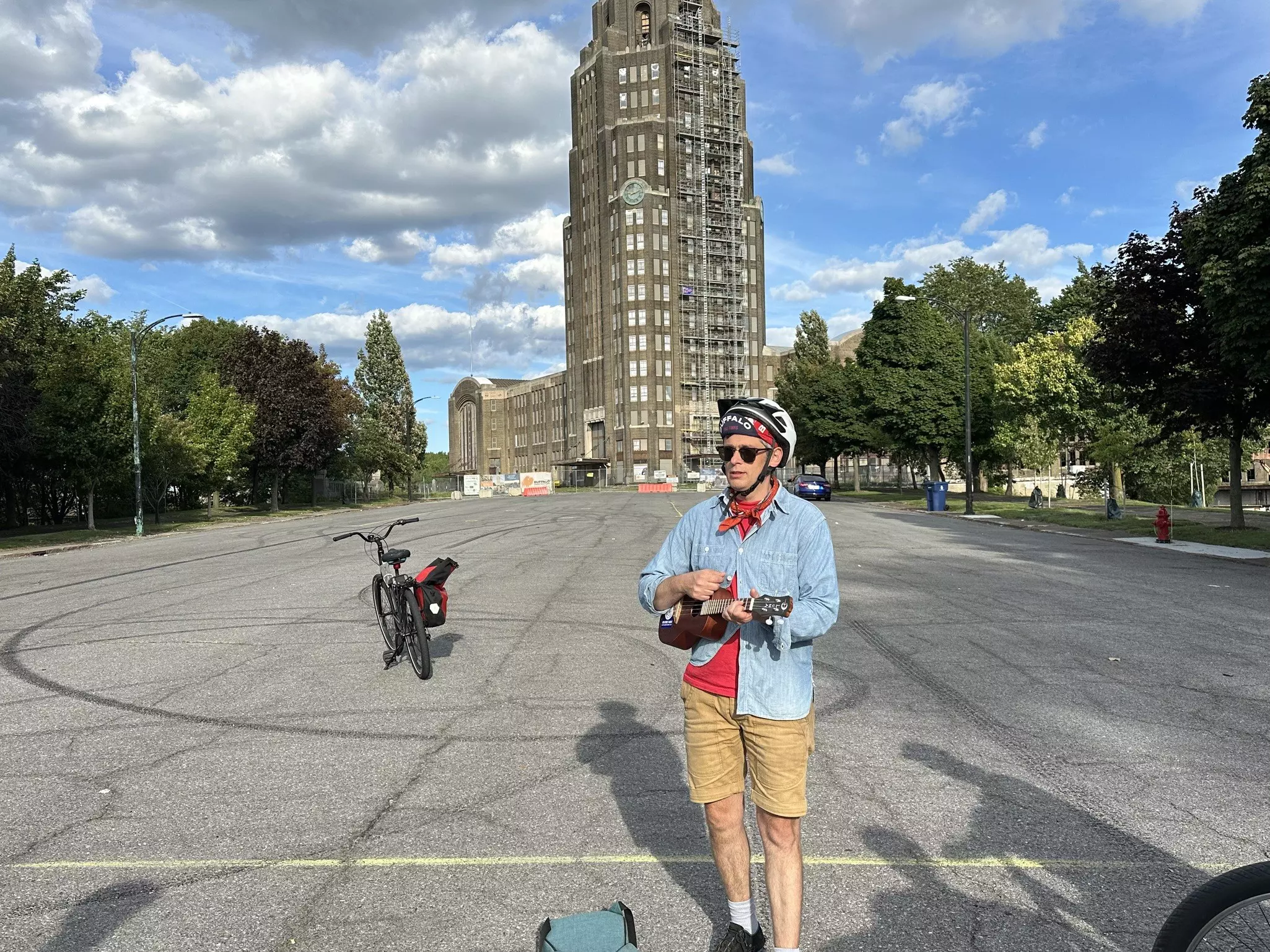 A man in shorts and blue top, holding a ukelele outside a building. His bike is in the background.
