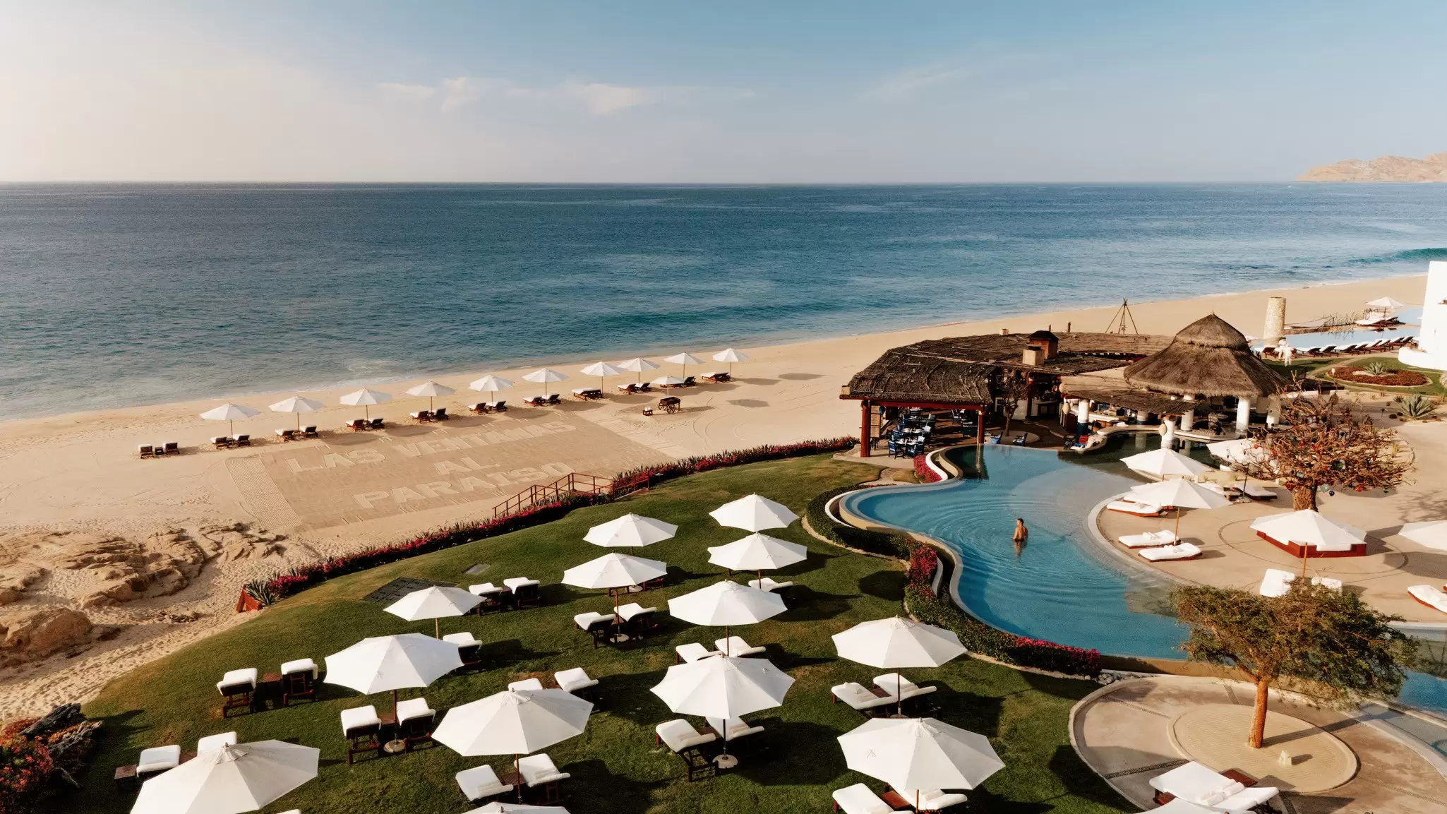 Exterior of a beachside hotel with a pool surrounded white lounge chairs covered by white umbrellas. A few of the same types of chairs and umbrellas line the beach