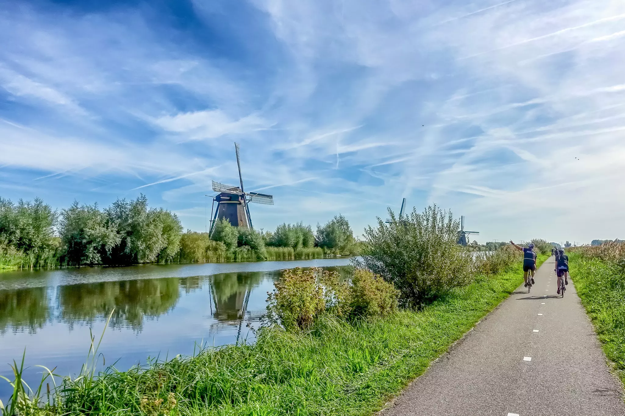 Cyclists ride bikes along a flat dedicated path beside a canal lined with windmills