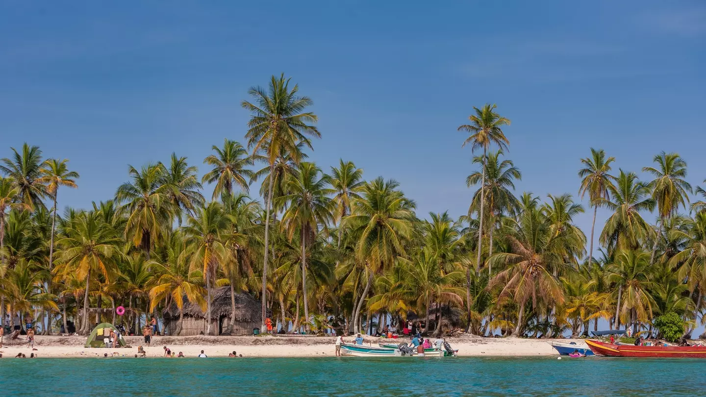 Palm trees back a white sand beach; a few boats are pulled up on the sand, and some people are in the blue water.