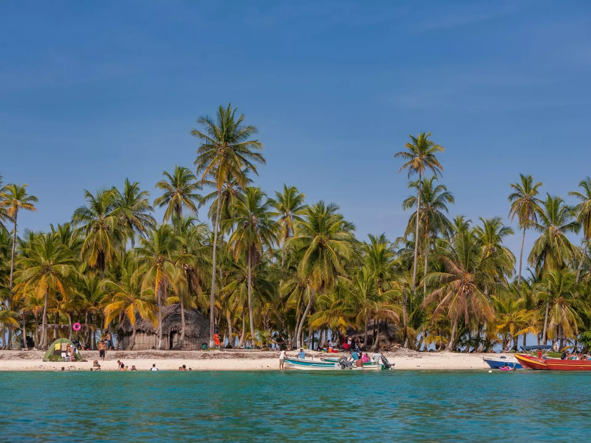 Palm trees back a white sand beach; a few boats are pulled up on the sand, and some people are in the blue water.