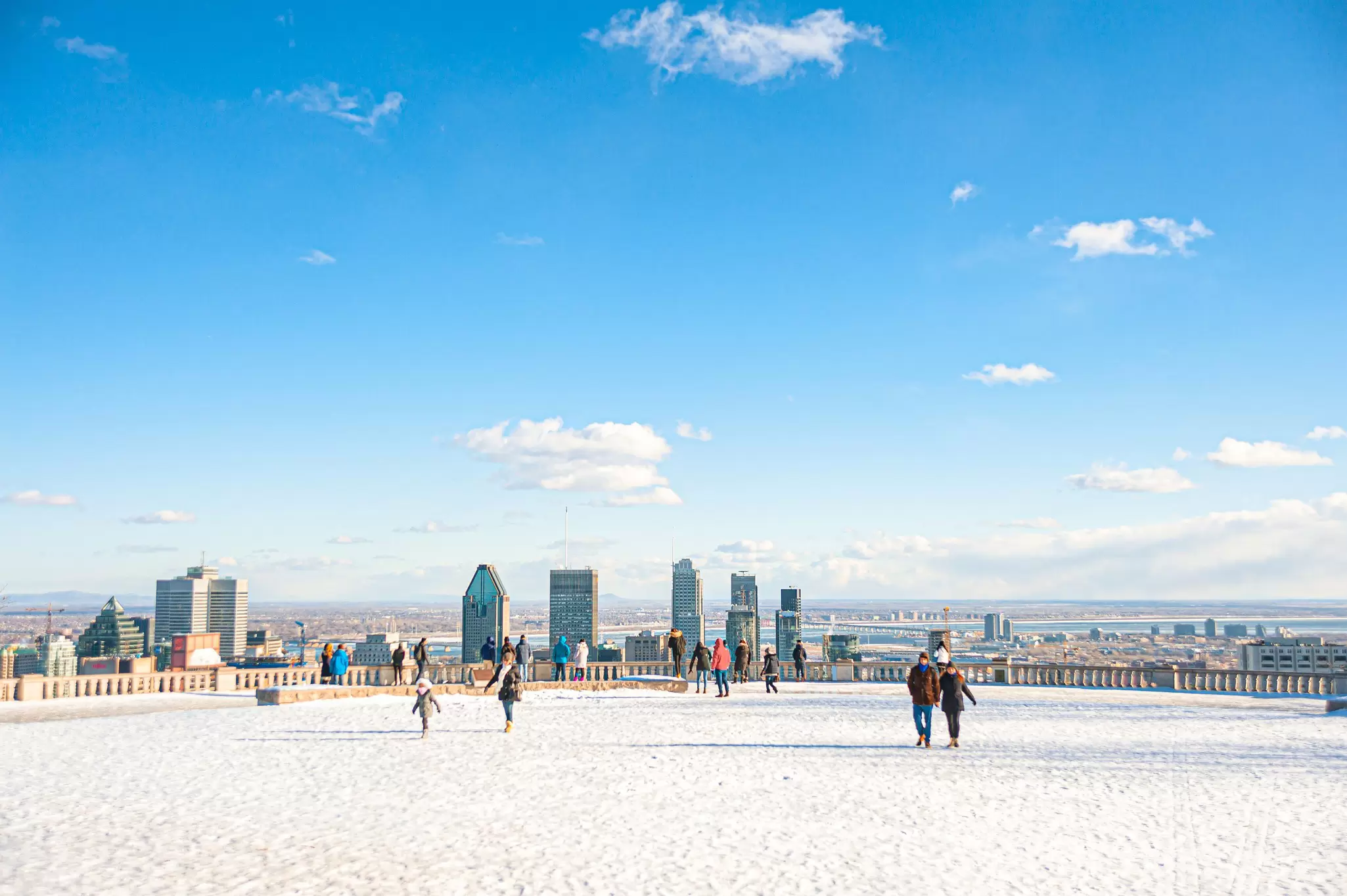 People wander over a snow-covered viewpoint in parkland that faces towards a city skyline