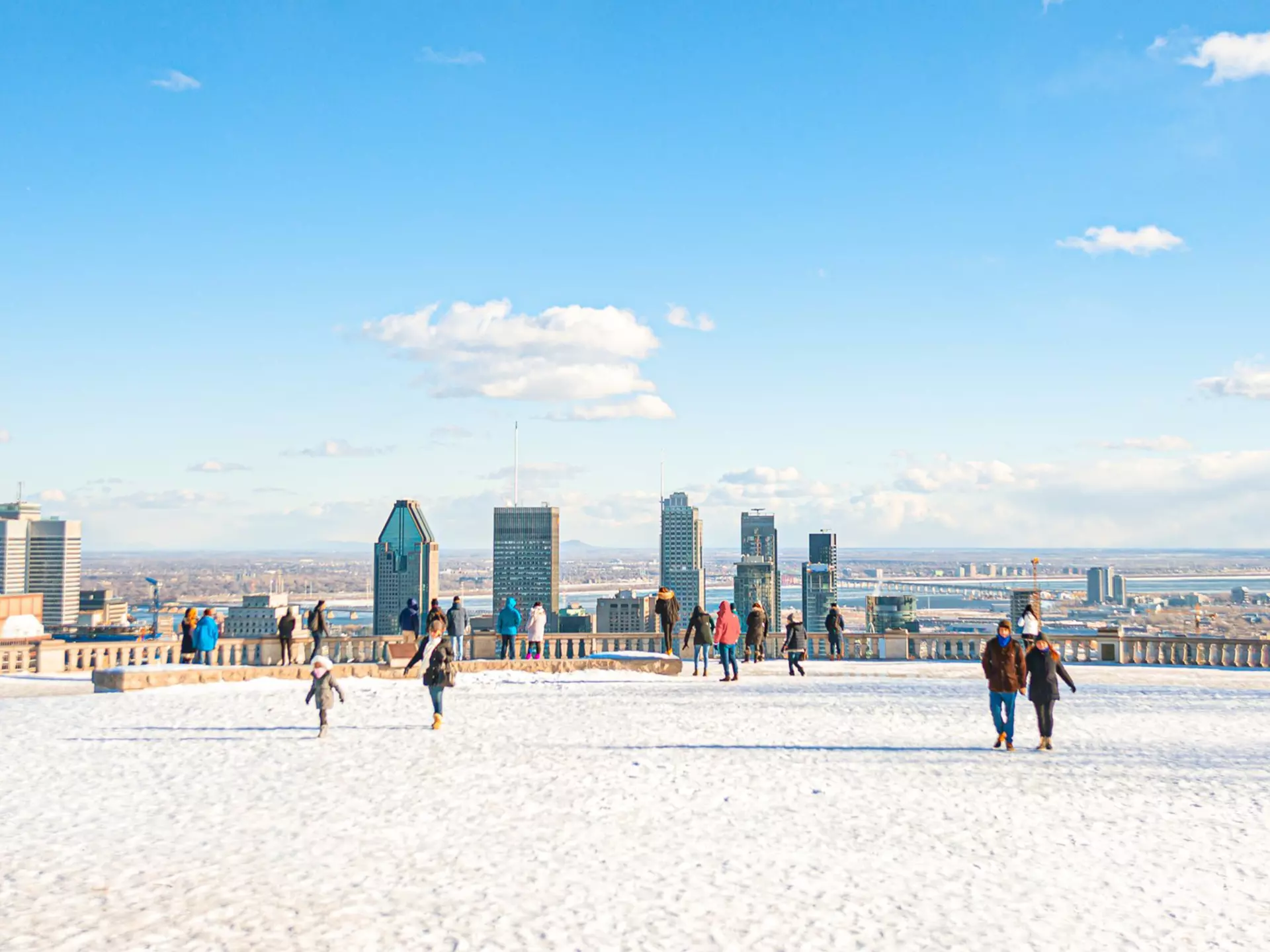 People wander over a snow-covered viewpoint in parkland that faces towards a city skyline