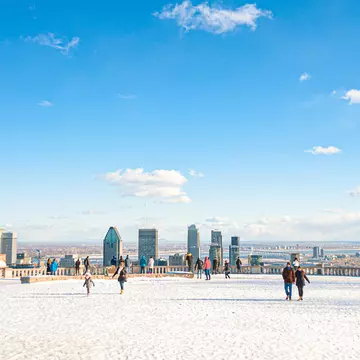 People wander over a snow-covered viewpoint in parkland that faces towards a city skyline