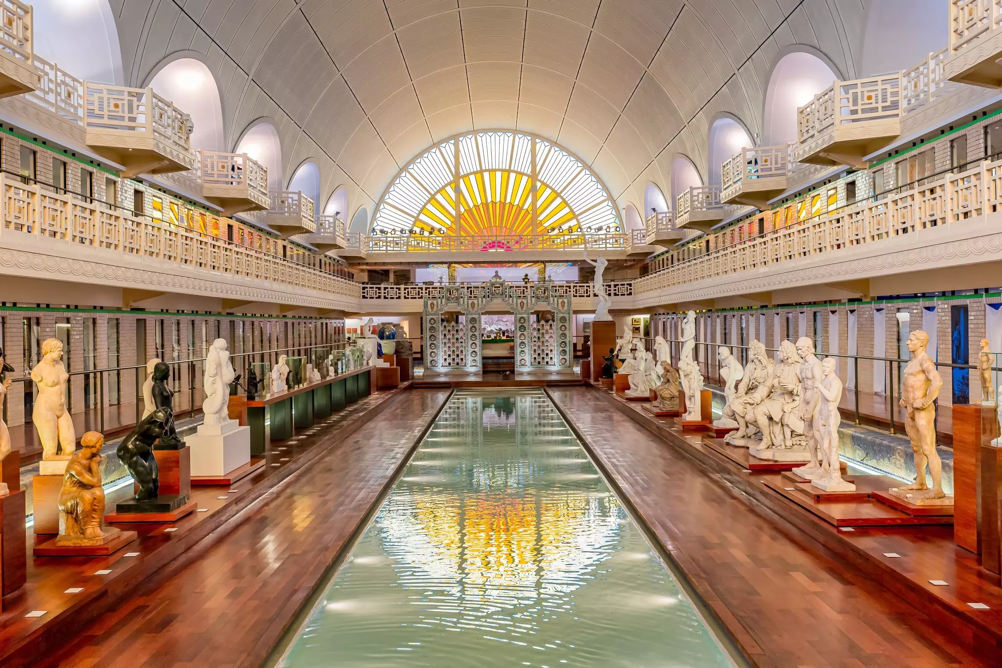 A long thin indoor swimming pool converted into a gallery space, with marble sculptures surrounding the pool and a large sun-like stained glass window reflecting in the water.