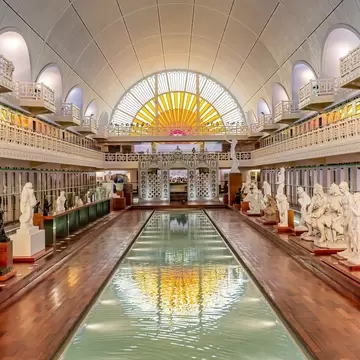 A long thin indoor swimming pool converted into a gallery space, with marble sculptures surrounding the pool and a large sun-like stained glass window reflecting in the water.