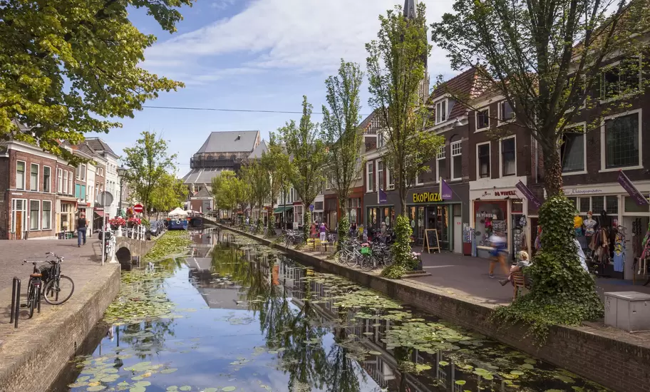 Delft canal covered in lily pads, lined with brick shops, bikes and trees, with a church spire behind