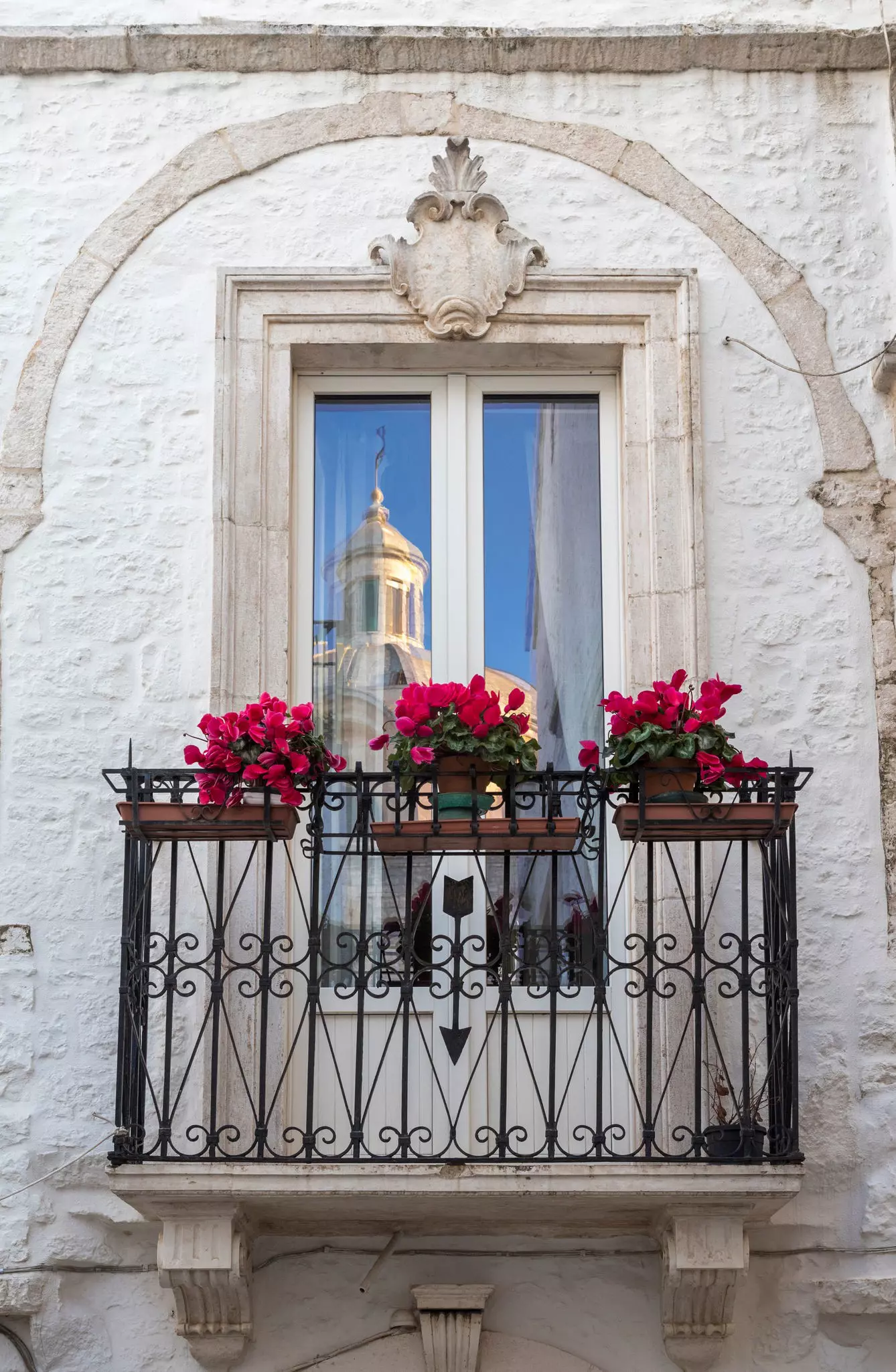 Balcony of a whitewashed house decorated with red geraniums in Locorotondo, Italy, with the dome of a church is reflected in the window