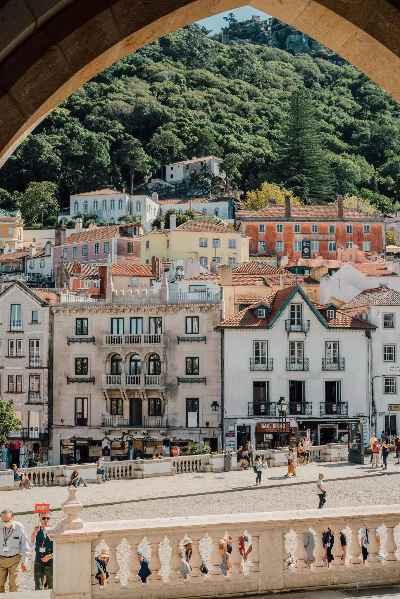 Pastel townhouses and a wooded hillside framed by a stone arch at the Sintra National Palace, with visitors below