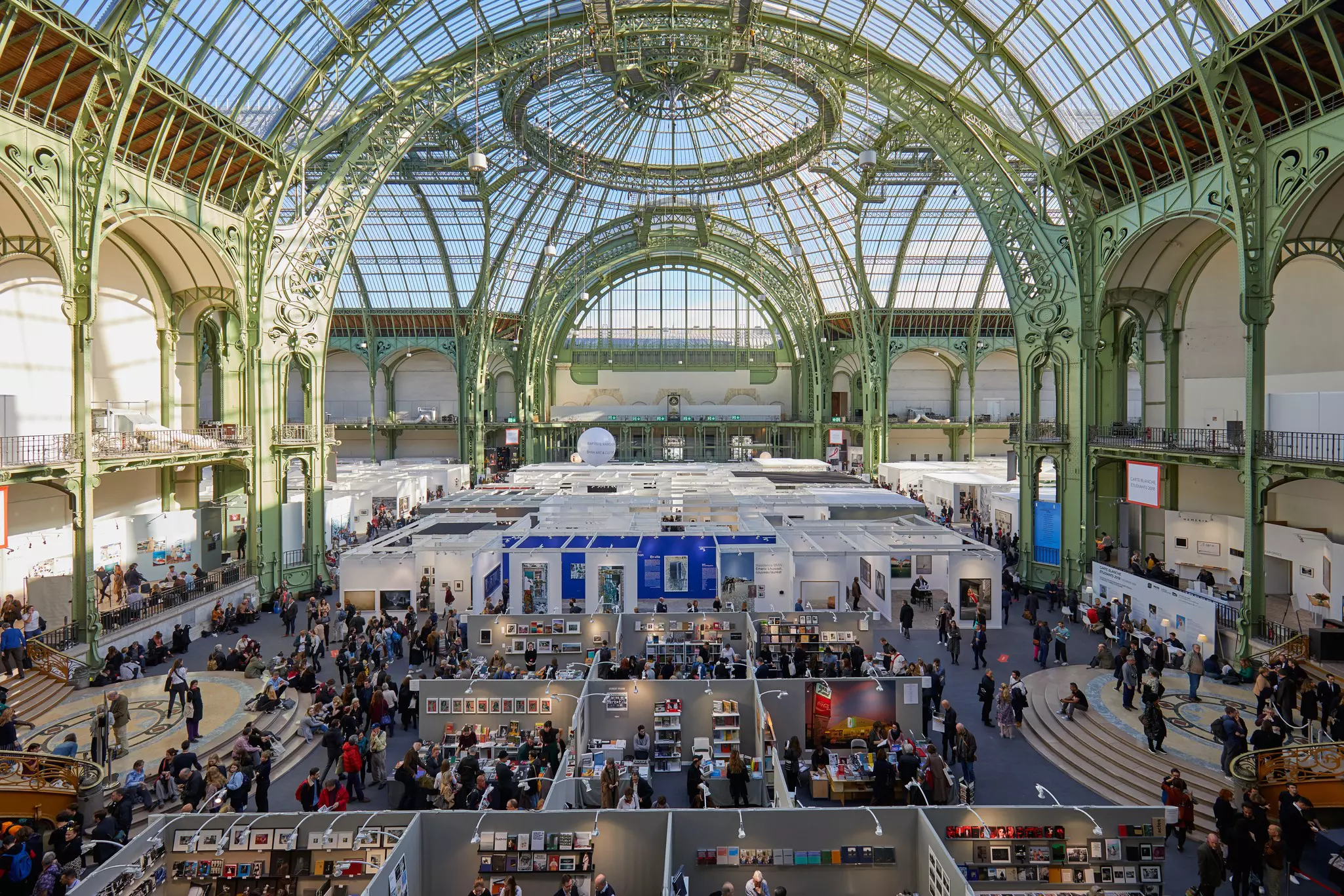 People gather for an exhibition in the main hall of the Grand Palais, featuring a striking iron, steel, and glass barrel-vaulted roof.