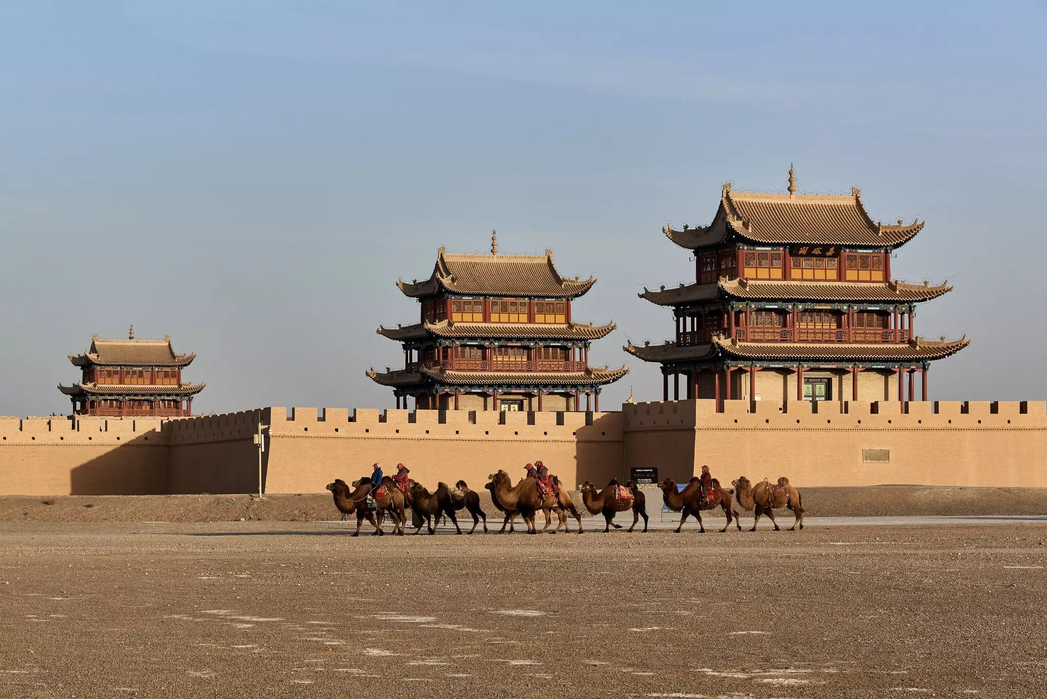 A caravan of camels crosses a sandy expanse in front of a wall, behind which three terraces towers rise.