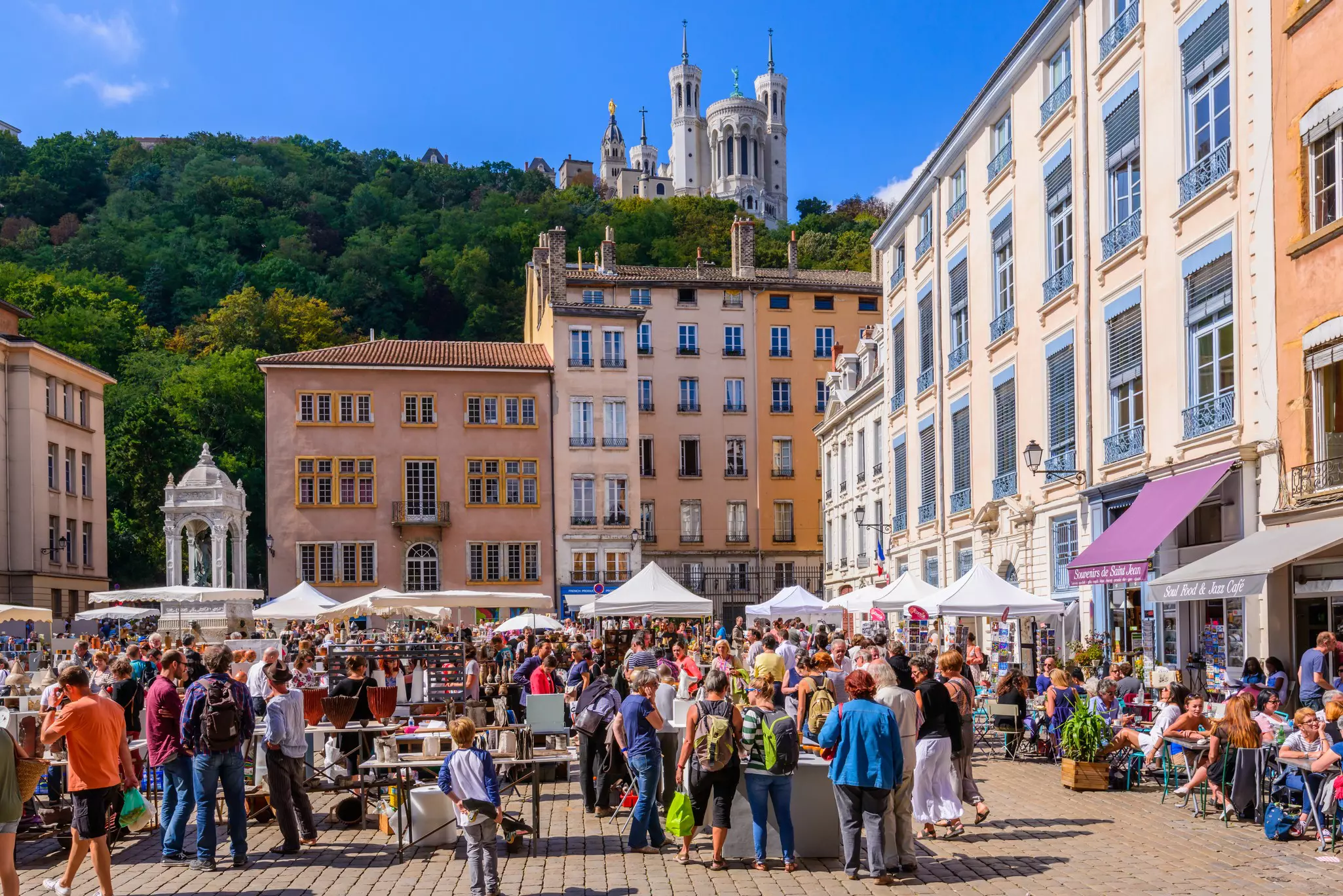 People shop at a market in a city square with a large white-stone cathedral on the hill above