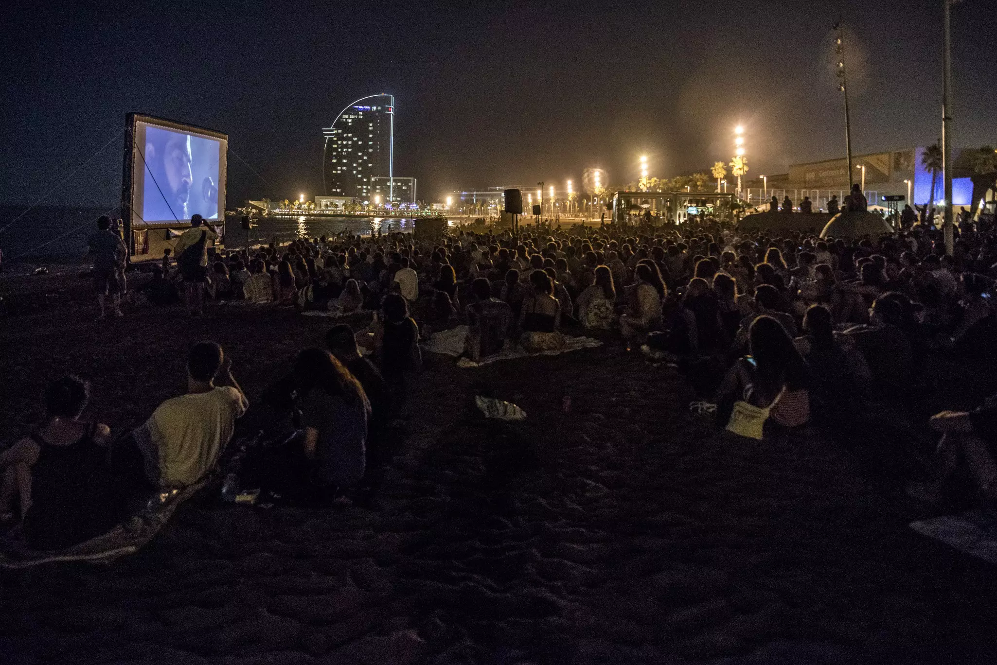 A crowd of people sitting on a beach in Barcelona at night watching a movie on a large screen.