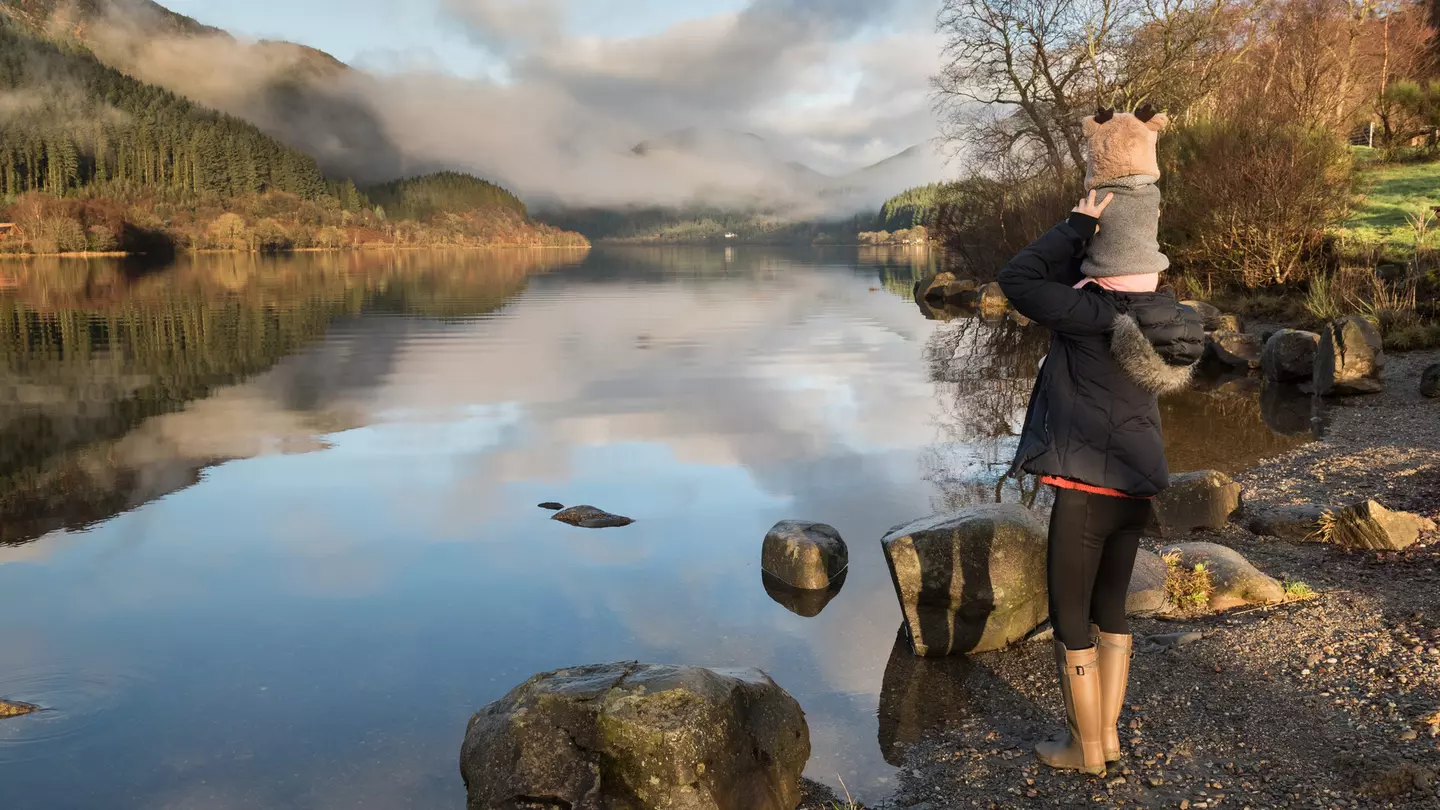Mother carrying baby on her shoulders glances at misty Loch Lomond in the Trossachs National Park, Scotland, UK