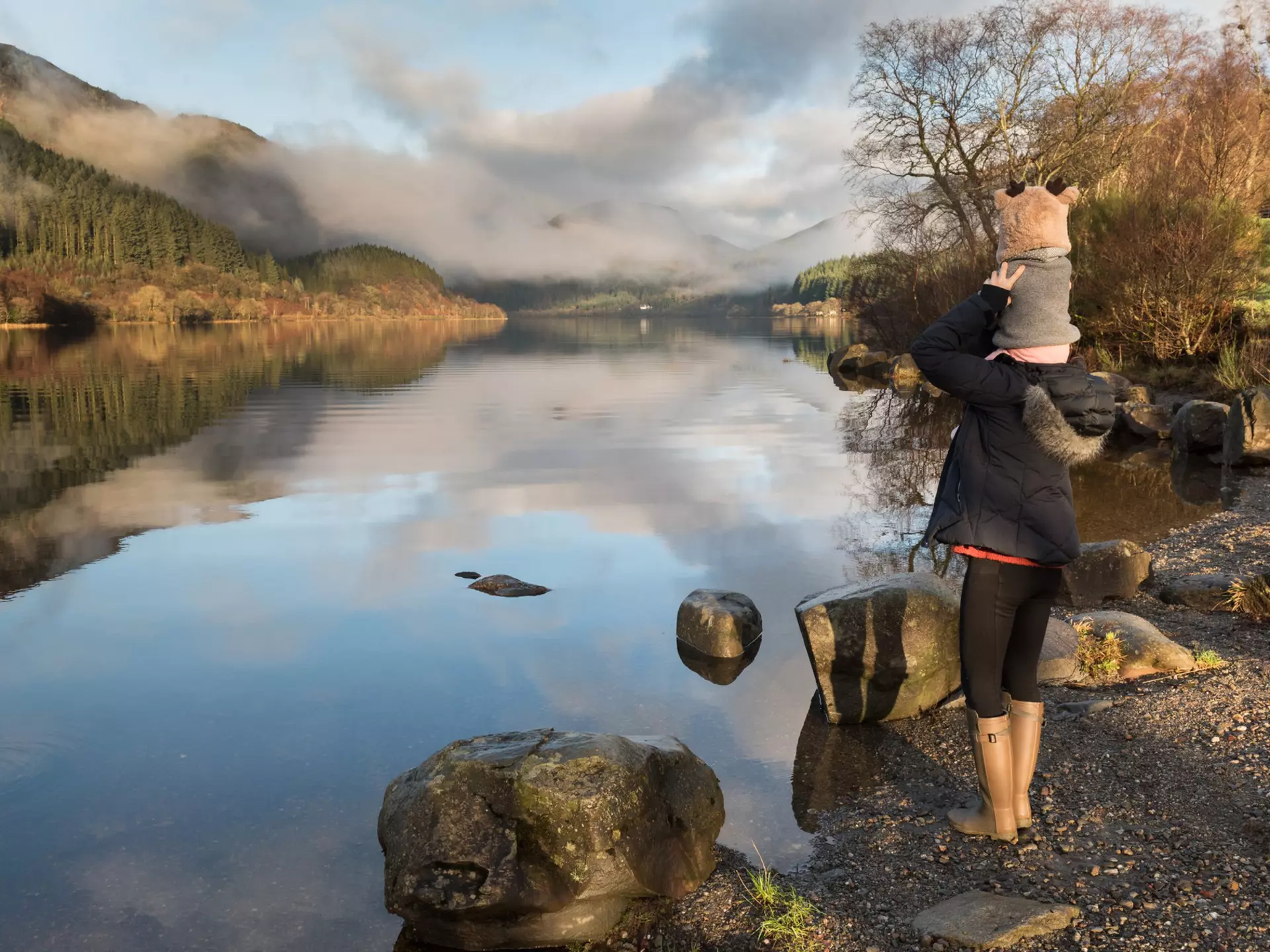Mother carrying baby on her shoulders glances at misty Loch Lomond in the Trossachs National Park, Scotland, UK