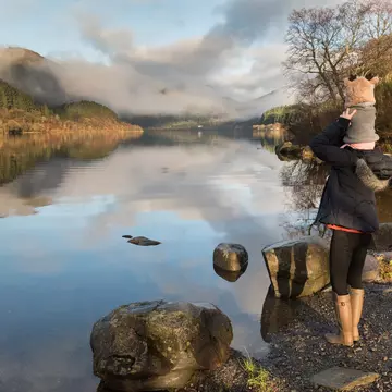 Mother carrying baby on her shoulders glances at misty Loch Lomond in the Trossachs National Park, Scotland, UK