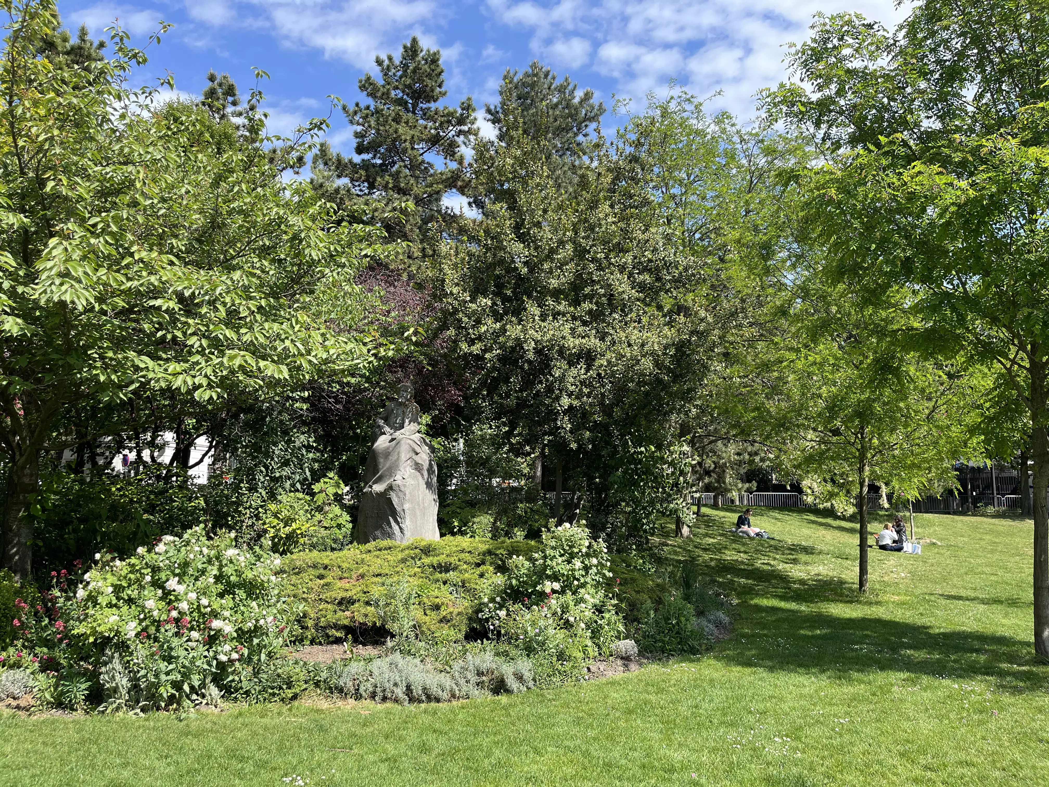 A peaceful green garden with sculptures dotted among the flower beds. People relax on the lawns.
