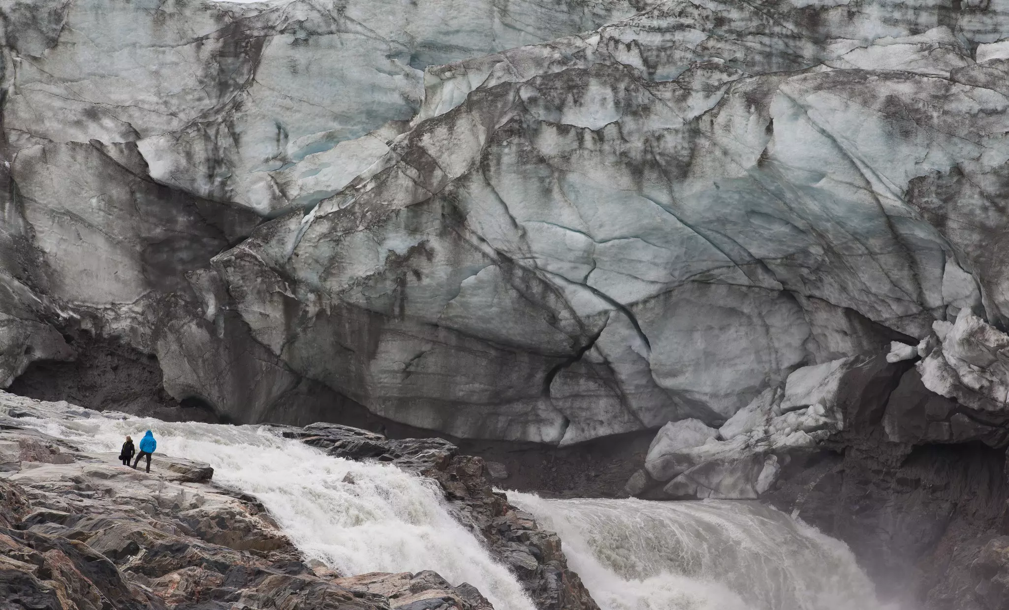 Two humans dwarfed by the vast craggy ice face covered in dark marks in front of them, as a river flows furiously by.
