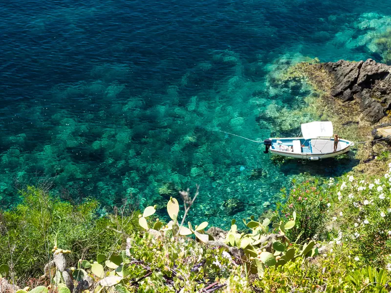 An overhead view of a small boat moored in a cove on an island. The water is many shades of blue, and green vegetation covers the slope leading down to the cove.
