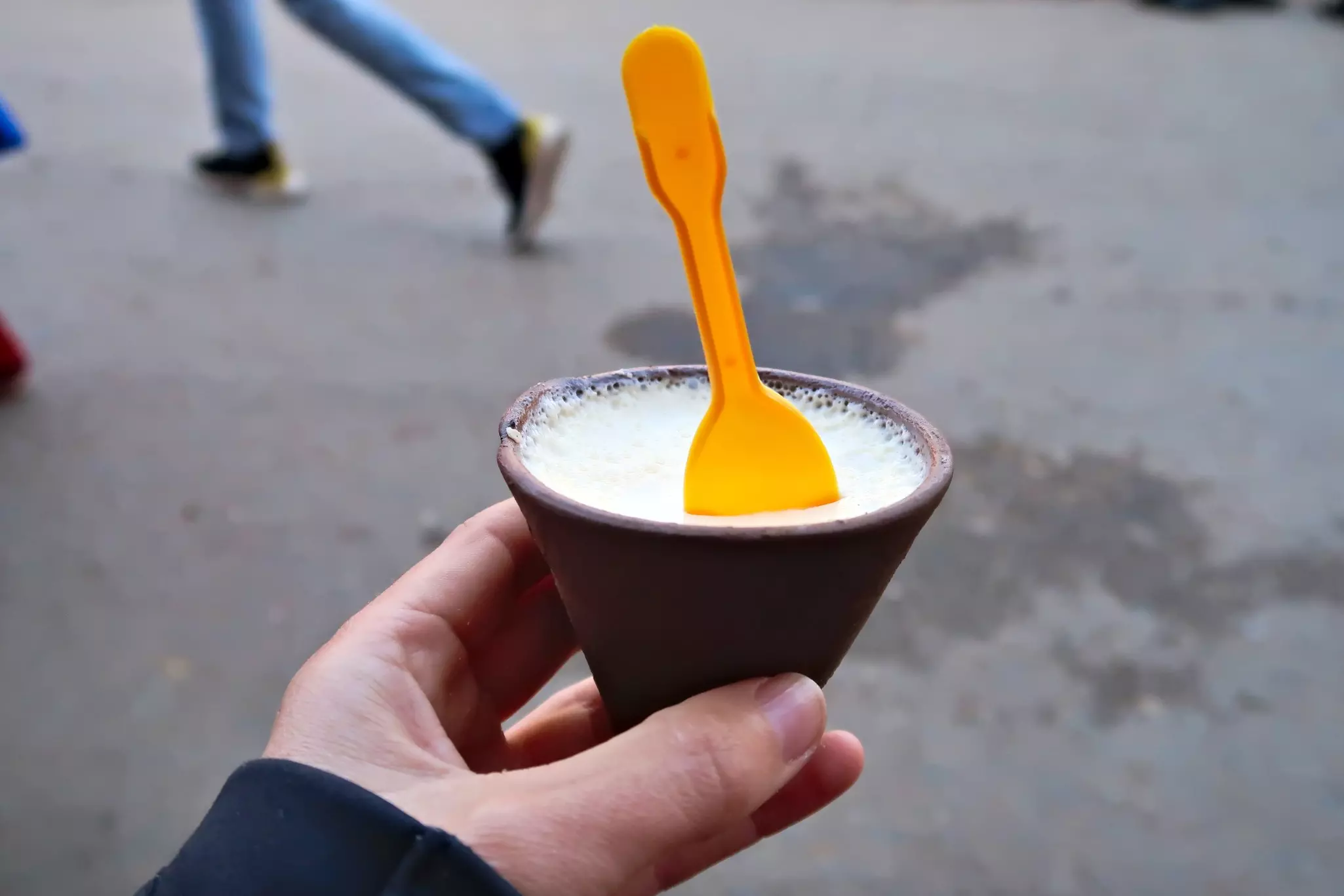 A hand holds a small, cone-shaped clay pot filled with yogurt. An orange spoon sticks out of the yogurt.