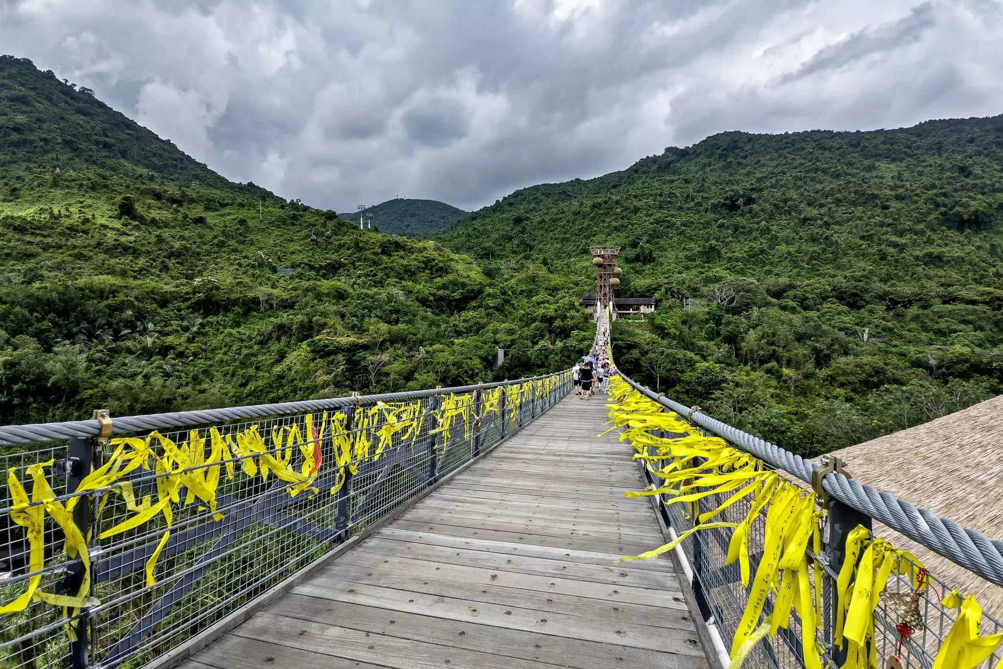 A pedestrian suspension adorned with yellow prayer flags spans a gorge in a tropical rainforest.