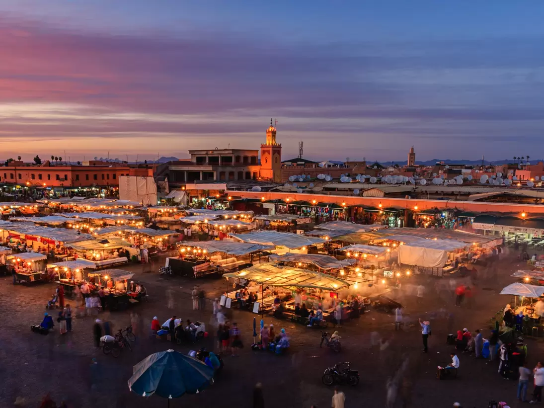 Aerial of a crowd at Djemaa El Fna square during the early evening in Marrakesh's Medina quarter; moving people are blurred between the umbrella-covered food stands. The mosque's minaret stands tall against the violet sky in the background.