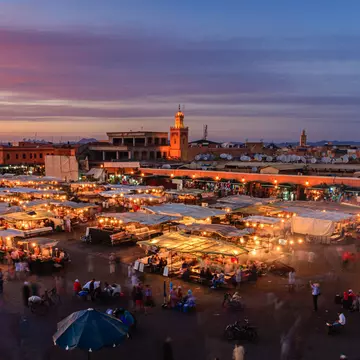 Aerial of a crowd at Djemaa El Fna square during the early evening in Marrakesh's Medina quarter; moving people are blurred between the umbrella-covered food stands. The mosque's minaret stands tall against the violet sky in the background.