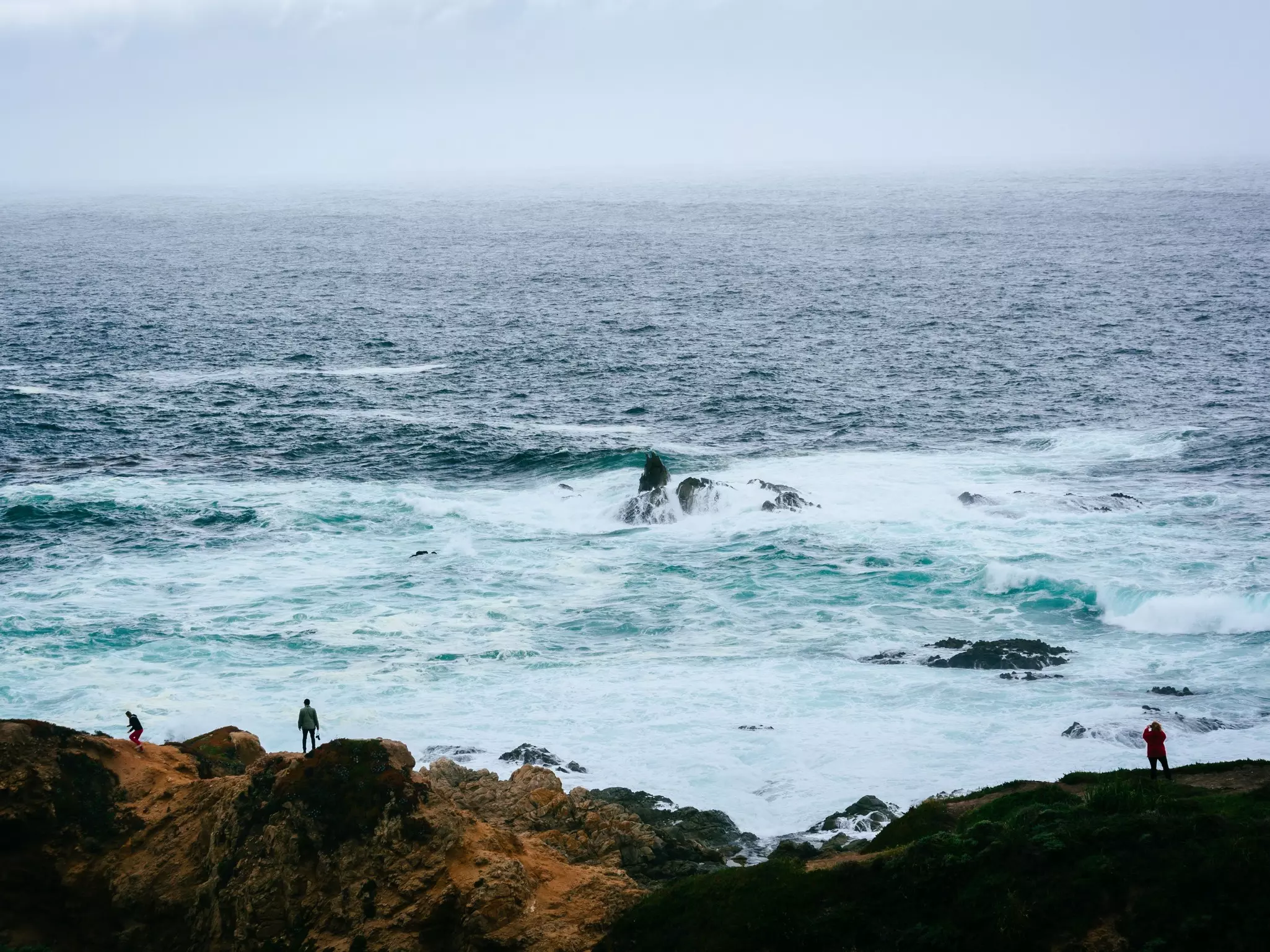 People stand on the rocky coastline looking out to sea where the waves are churning into white froth.
