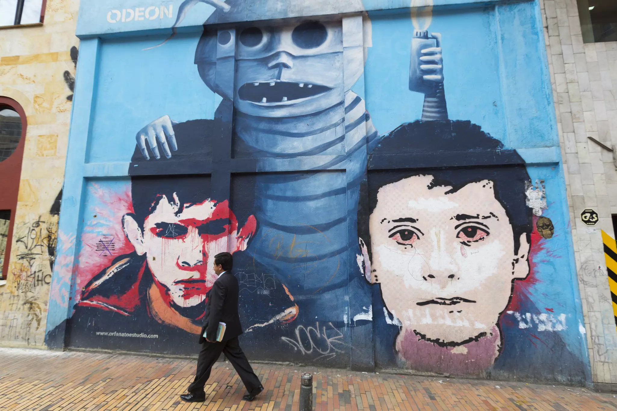 A man walks by a street mural on a city street. The mural depicts two boys’ faces and a mythical creature.