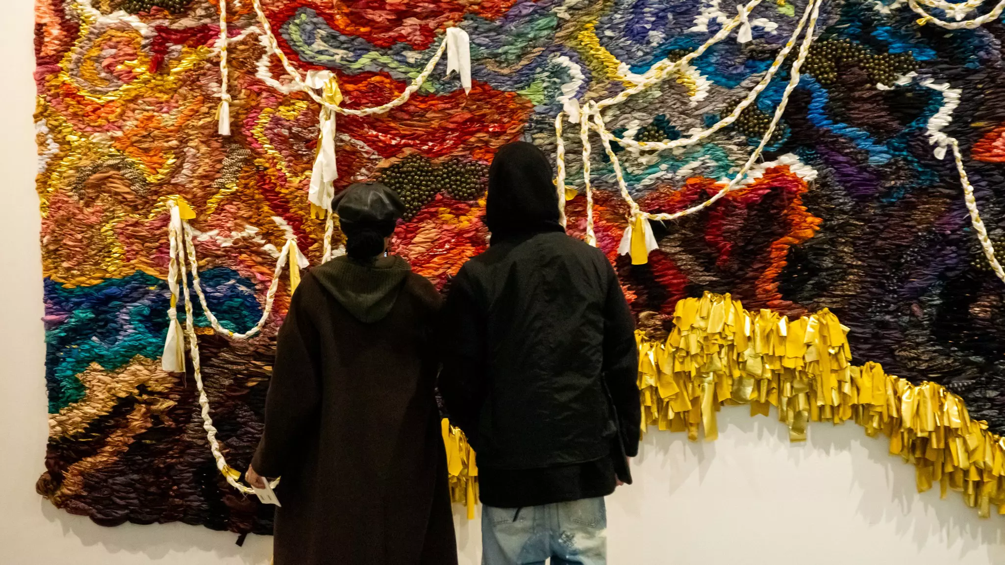 Two people are pictured from behind looking at an artwork made from colorful textiles and other materials.
