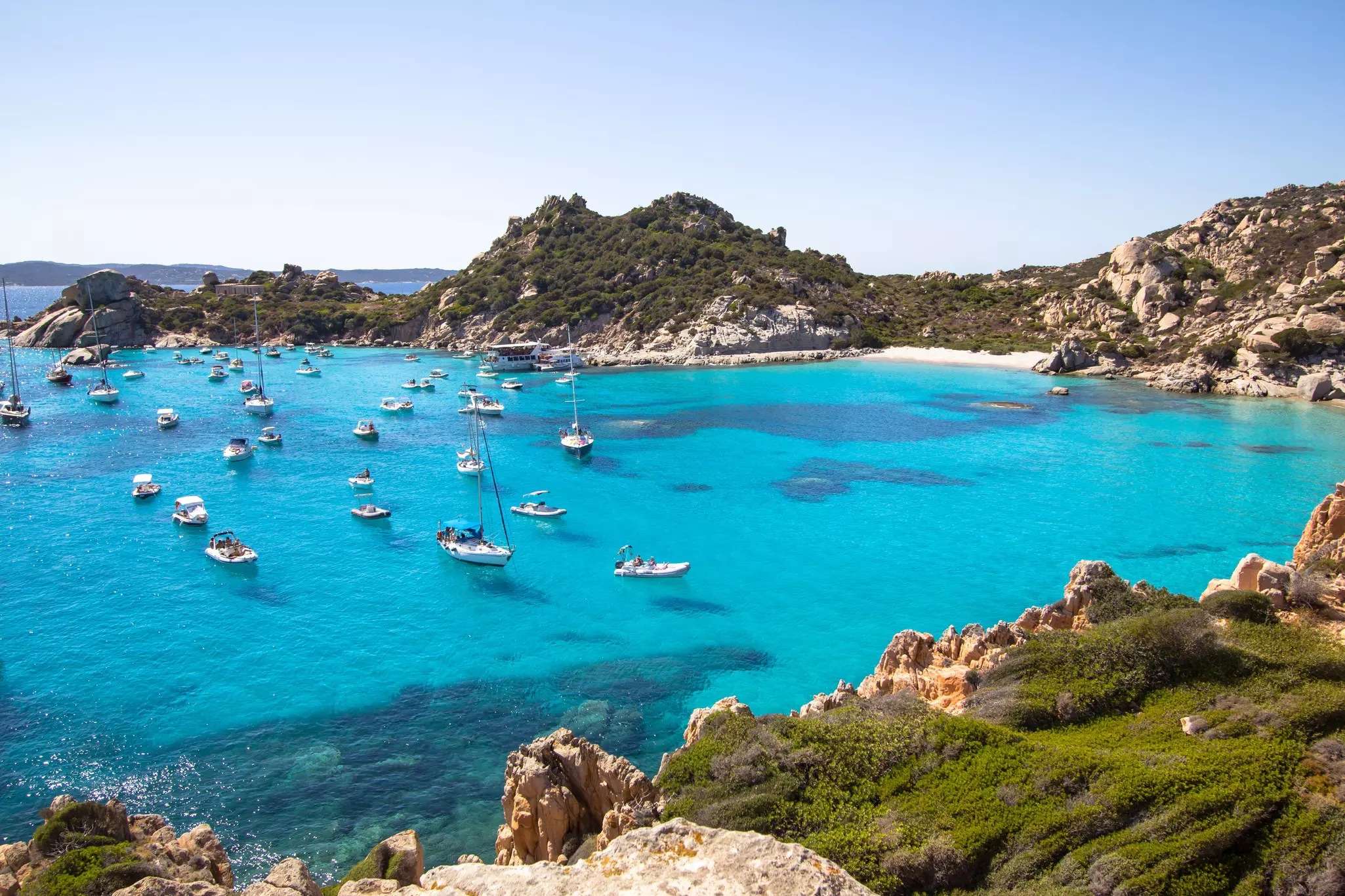Aerial view of many boats moored in clear, turquoise water surrounded by small islands on a sunny day.