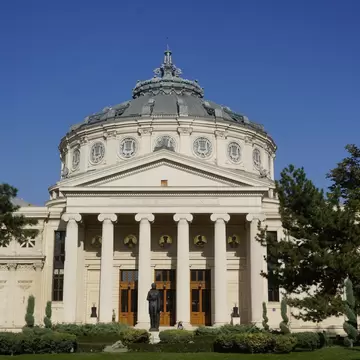 White neoclassical hall with Ionic columns and a grey dome, a bronze statue in the garden in front.