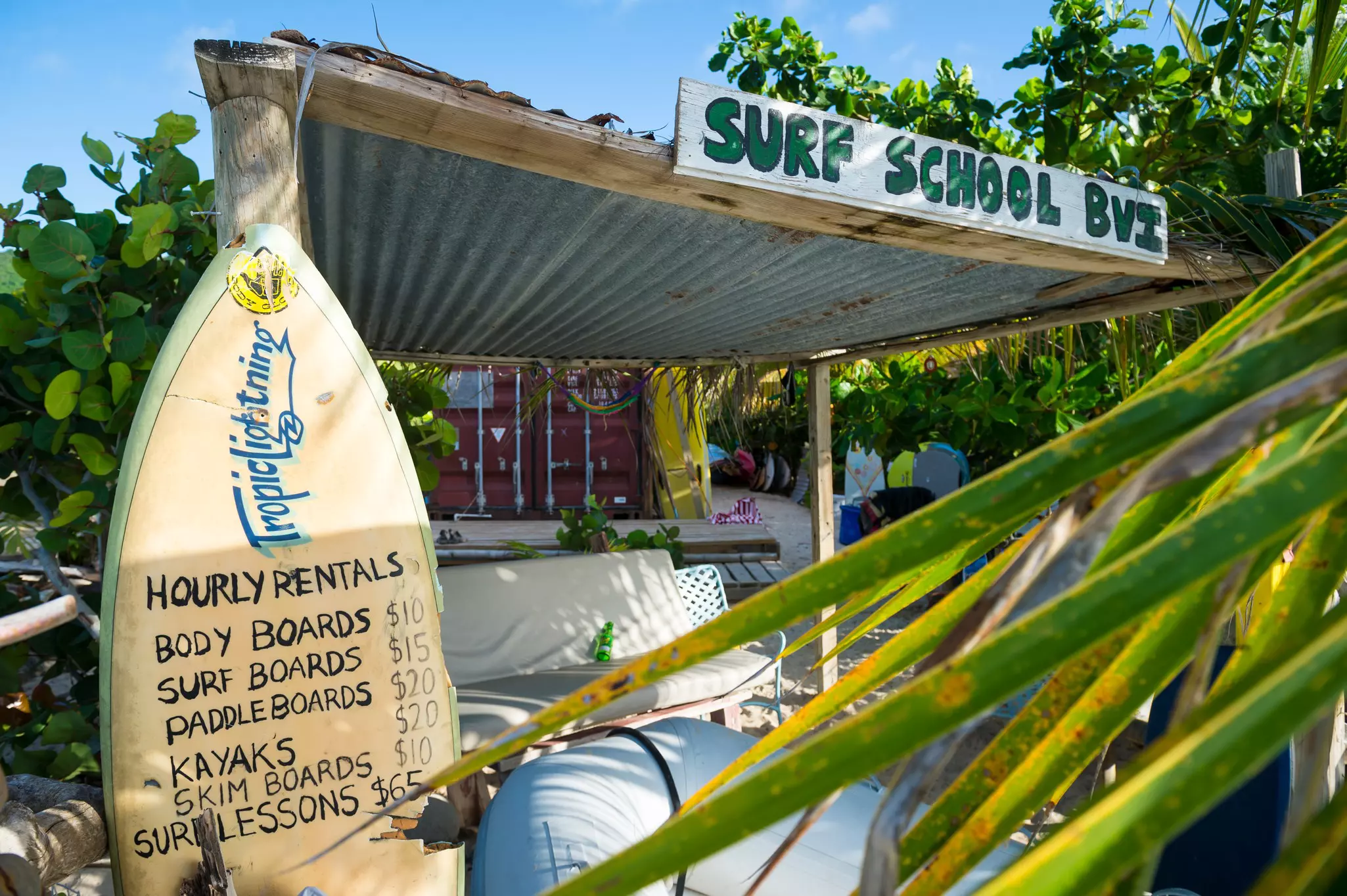 A surfboard displays prices at a beachfront surf school surrounded by lush greenery in a tropical destination.