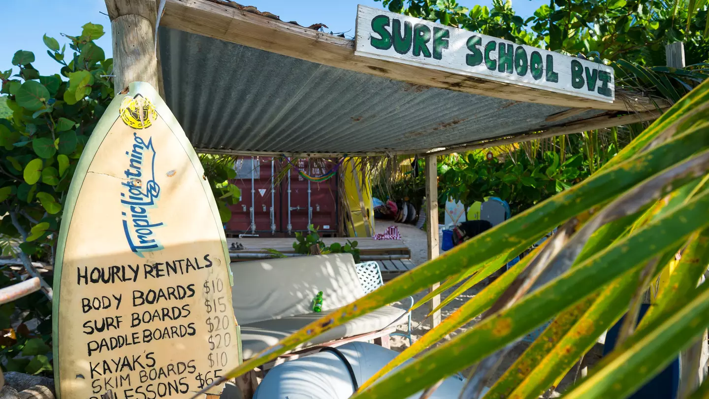 A surfboard displays prices at a beachfront surf school surrounded by lush greenery in a tropical destination.