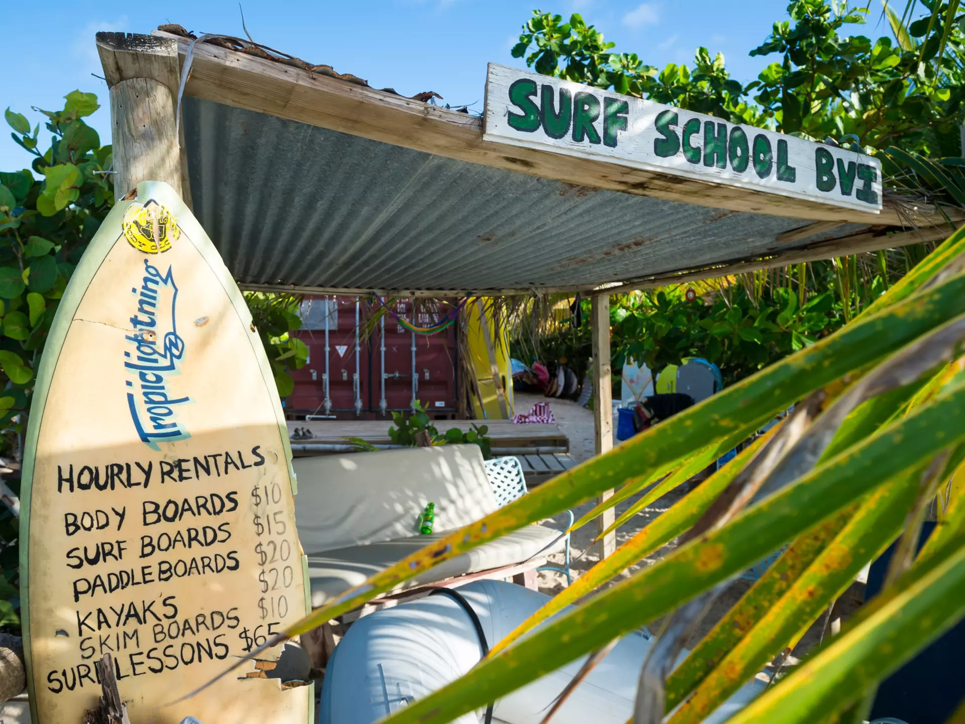 A surfboard displays prices at a beachfront surf school surrounded by lush greenery in a tropical destination.