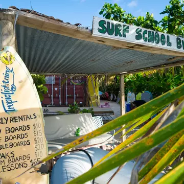 A surfboard displays prices at a beachfront surf school surrounded by lush greenery in a tropical destination.