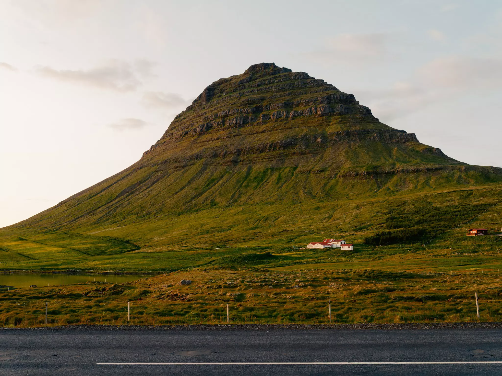 Grass-covered mountain with a cluster of white houses with red roofs at its base, another reddish house to the right and a roadway in the foreground on an overcast day.