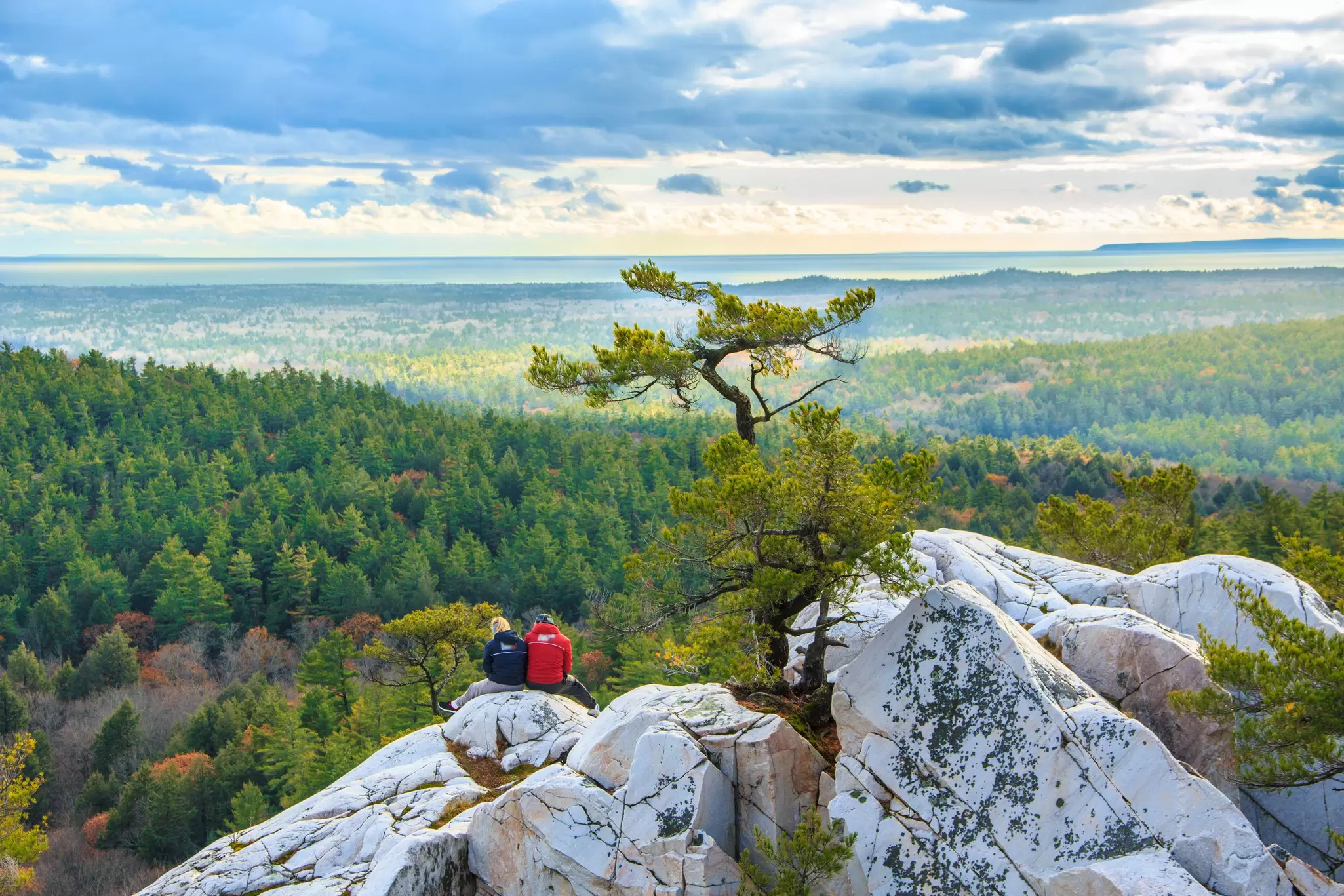 Two people sit on a rocky outcrop looking out over a panoramic view of pine trees, Killarney Provincial Park, Ontario