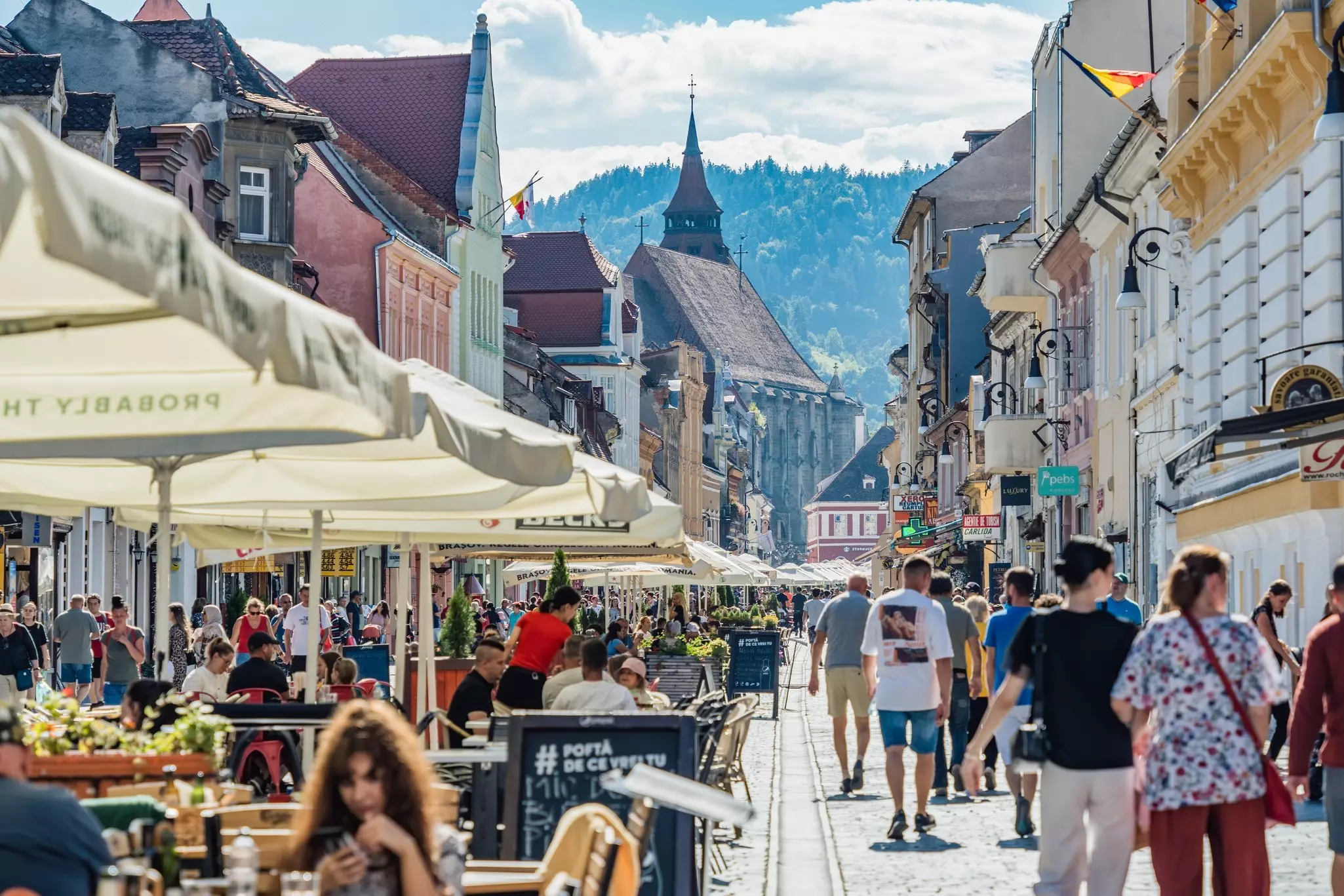 People walk through the pedestrian area in the old town center of Brasov, lined with historic buildings, with forested hills in the distance.