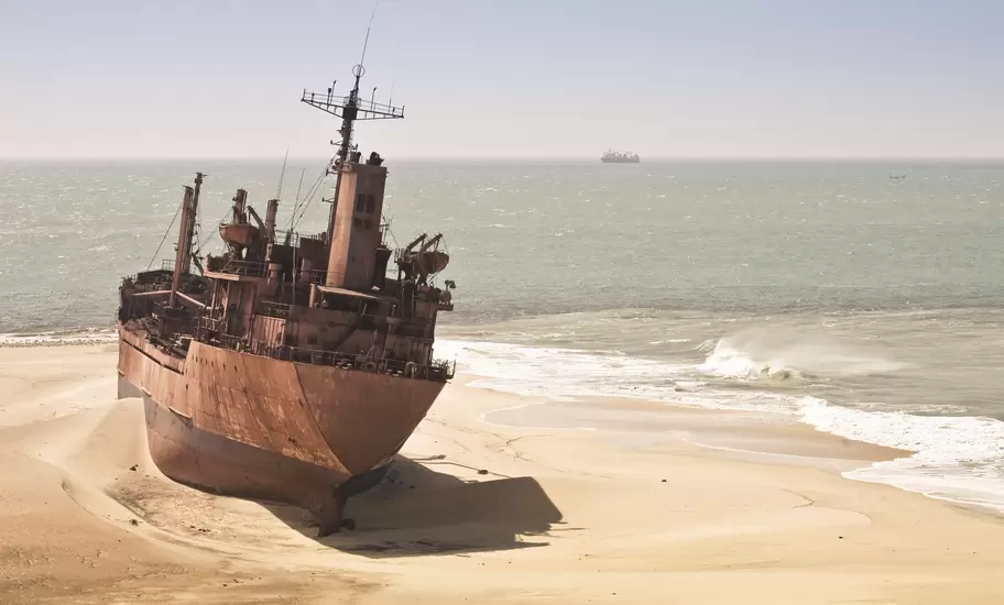 Rusting hull of a cargo ship beached on sand as waves break, Atlantic coast of Mauritania, another ship on the horizon