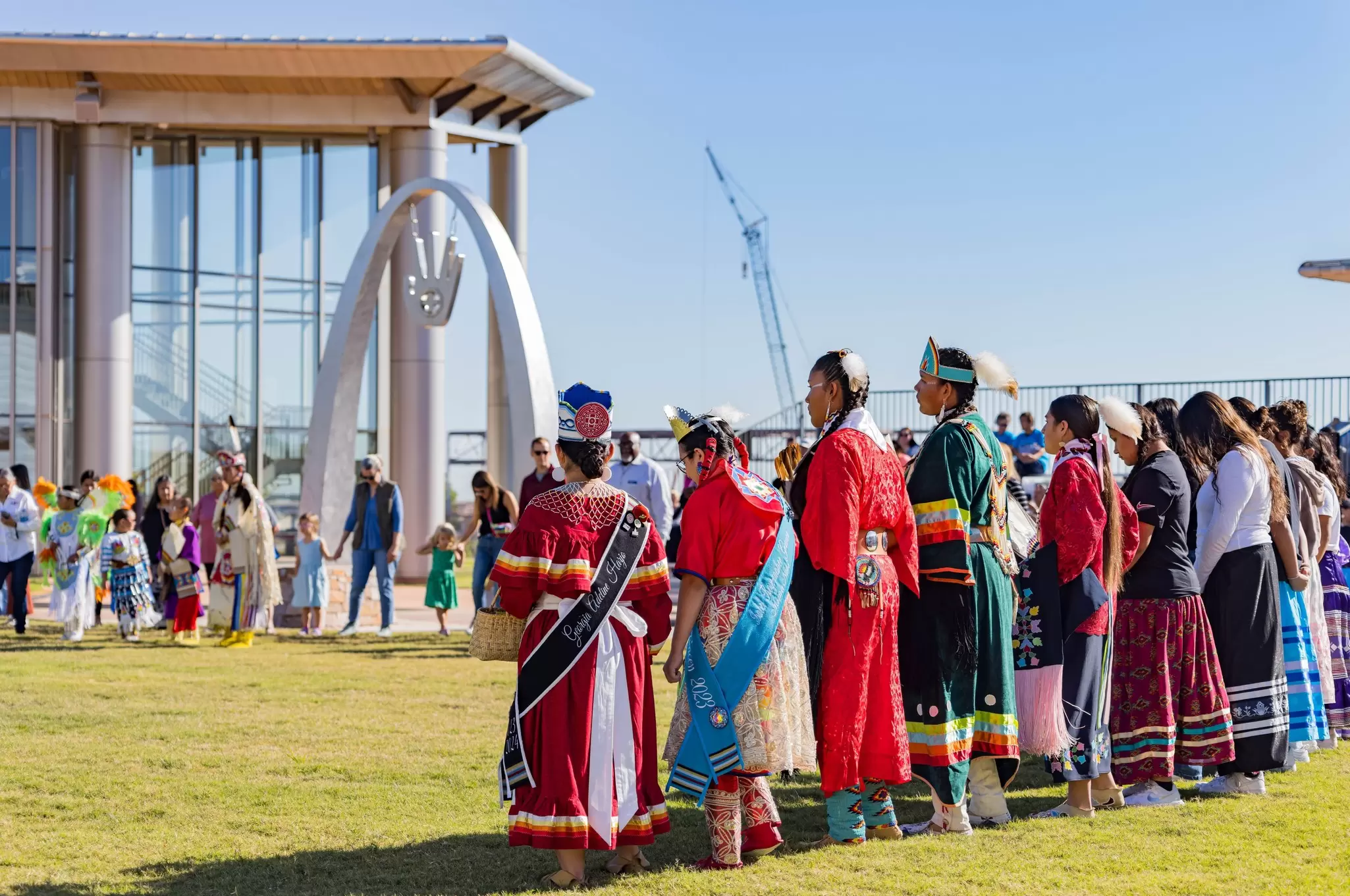A group of people in different styles and types of Indigenous clothing stand together in the grounds of a museum.