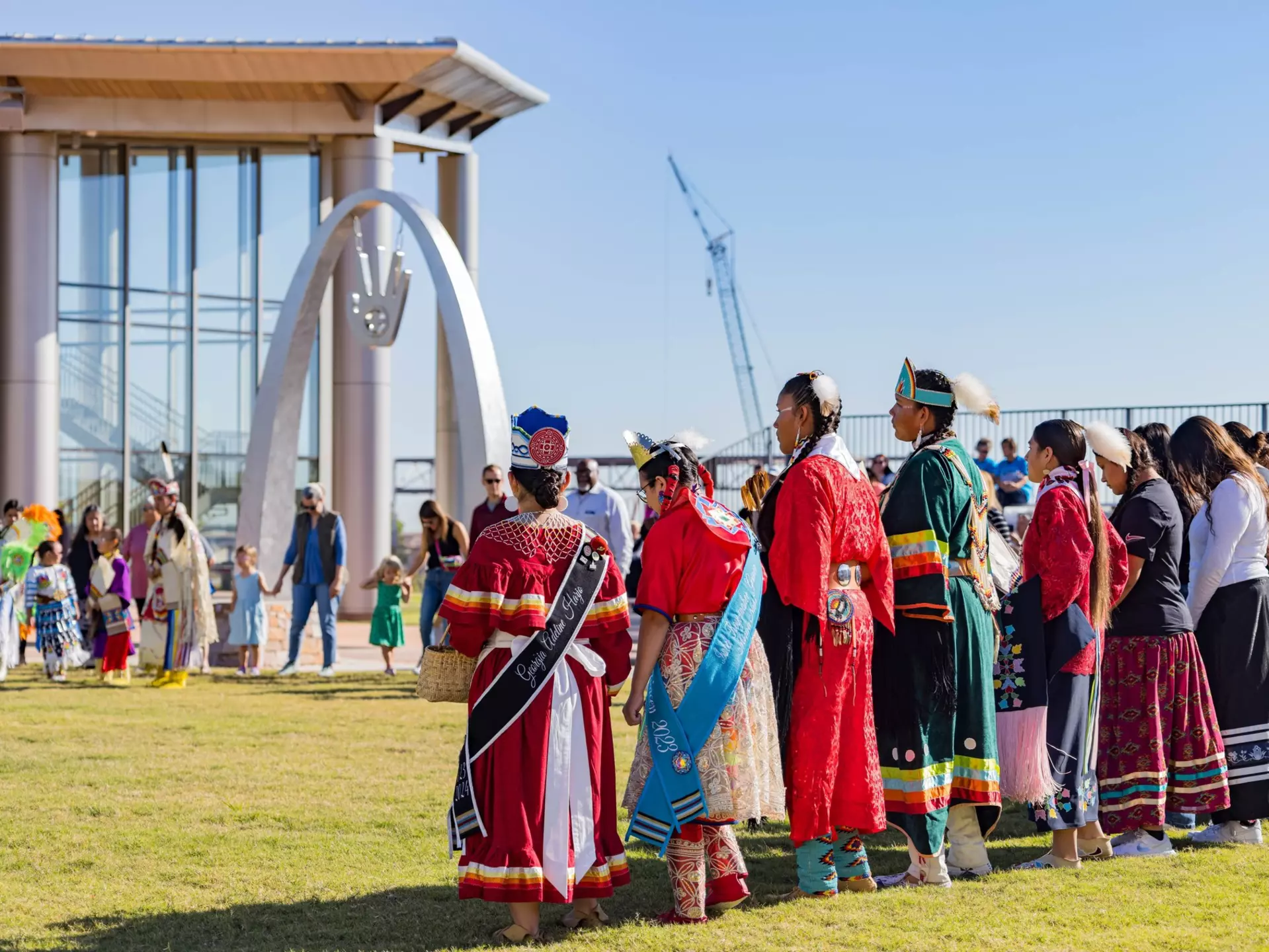 A group of people in different styles and types of Indigenous clothing stand together in the grounds of a museum.