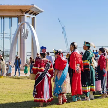 A group of people in different styles and types of Indigenous clothing stand together in the grounds of a museum.