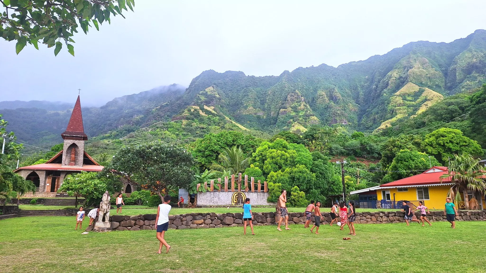 A group of young children play soccer on verdant grass with equally verdant mountains, an earth-toned church and a yellow house with a red roof in the background