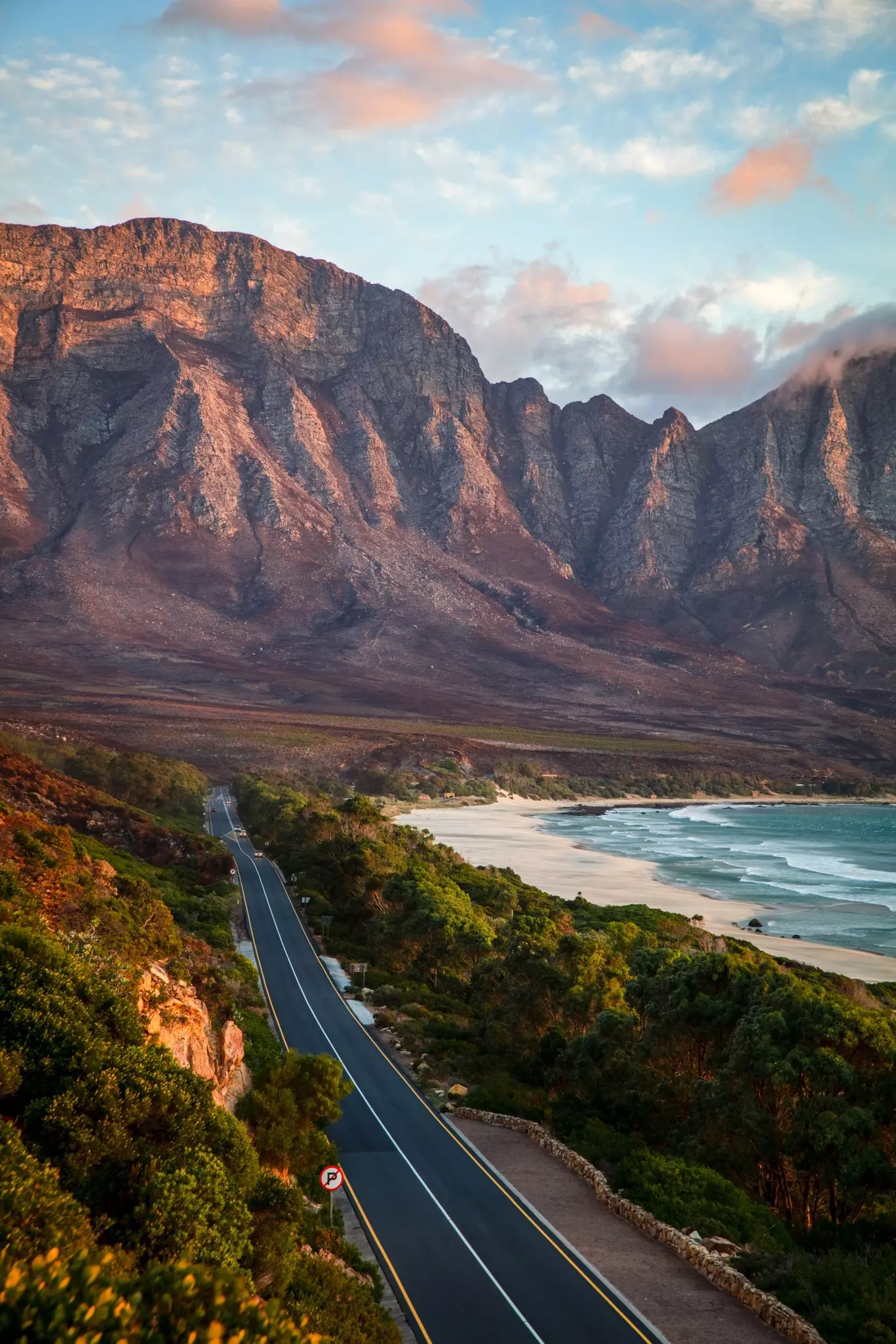 Two-lane road surrounded by green trees with ocean to the right and reddish rocky mountains in the distance at sunset.