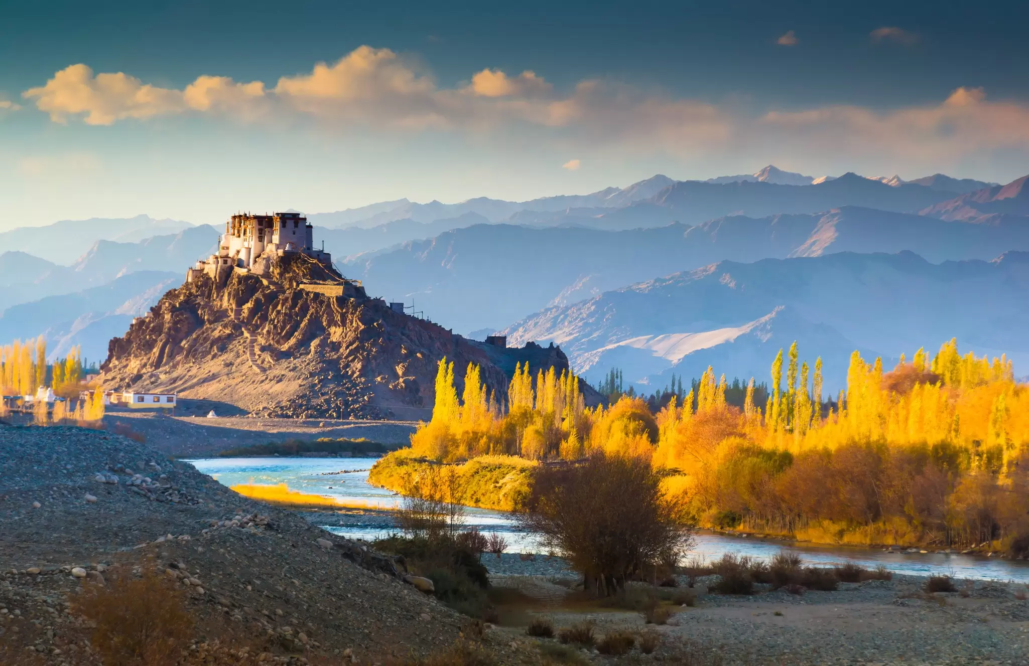 The sun illuminates trees in yellow fall foliage next to a river and a massif topped with a temple, Ladakh, India