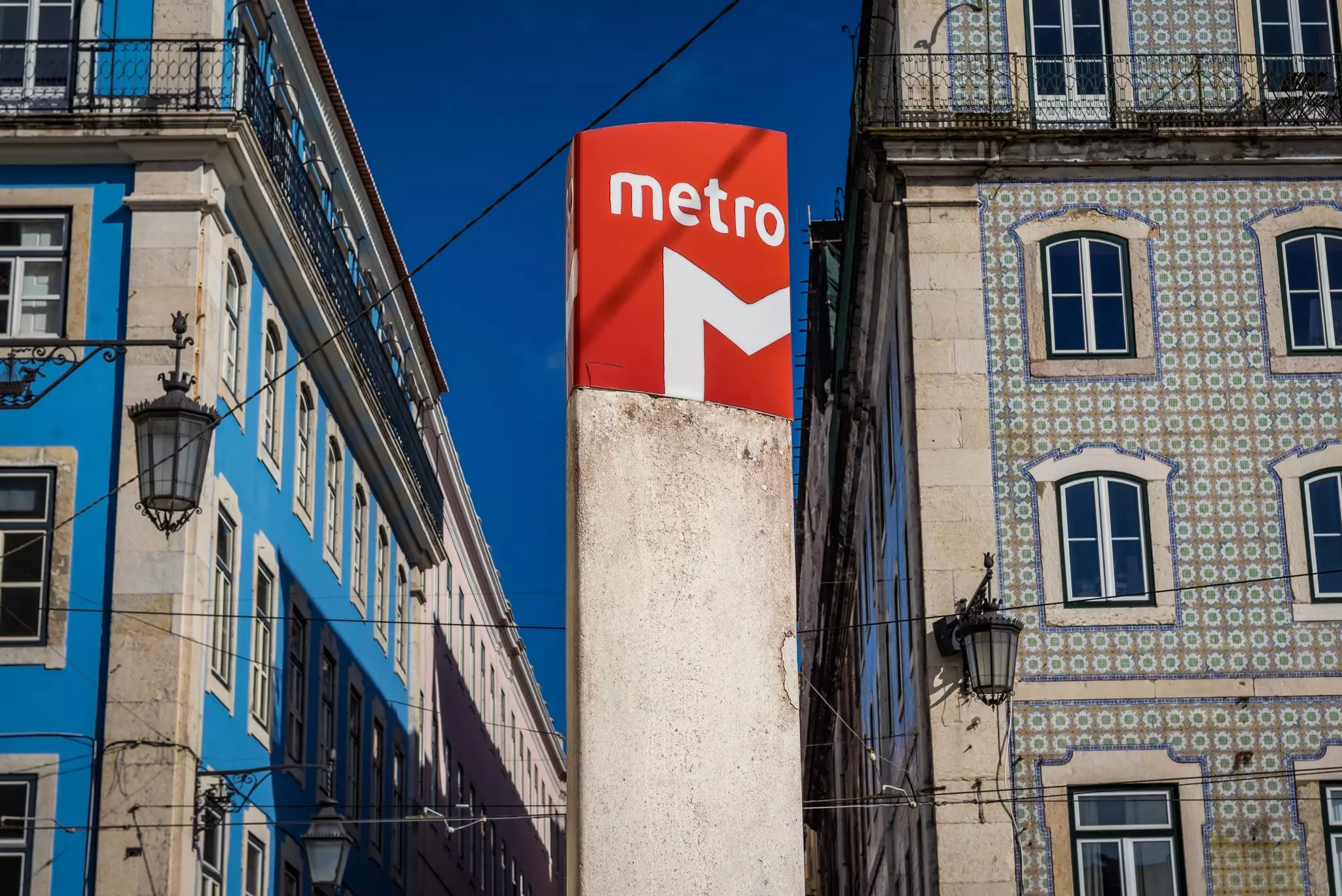 Tall cement post with red at top and "metro" and half of an "M" in white on a sunny day. There are two old buildings, blue on the left and tiles on the right.